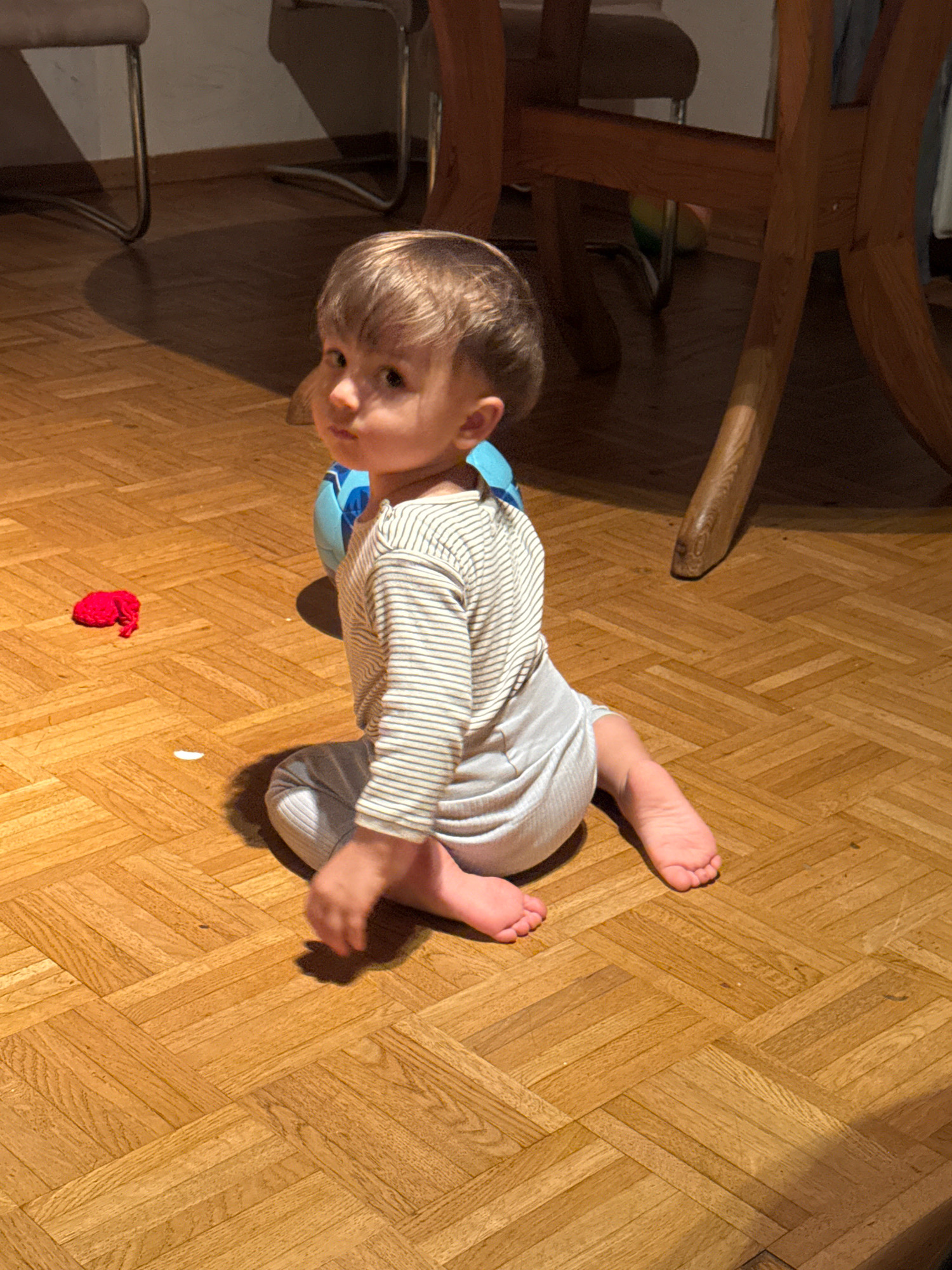 A toddler with blond hair sits on a parquet floor, looking over their shoulder. They are wearing a long-sleeved striped shirt and light-colored pants. A blue ball is partially visible behind them. To the left, on the floor, is a small red object. The scene appears to be indoors, likely a home, in Überherrn, Germany. The lighting suggests it may be evening, with shadows cast by furniture. The floor is made of wooden parquet laid in a herringbone pattern.