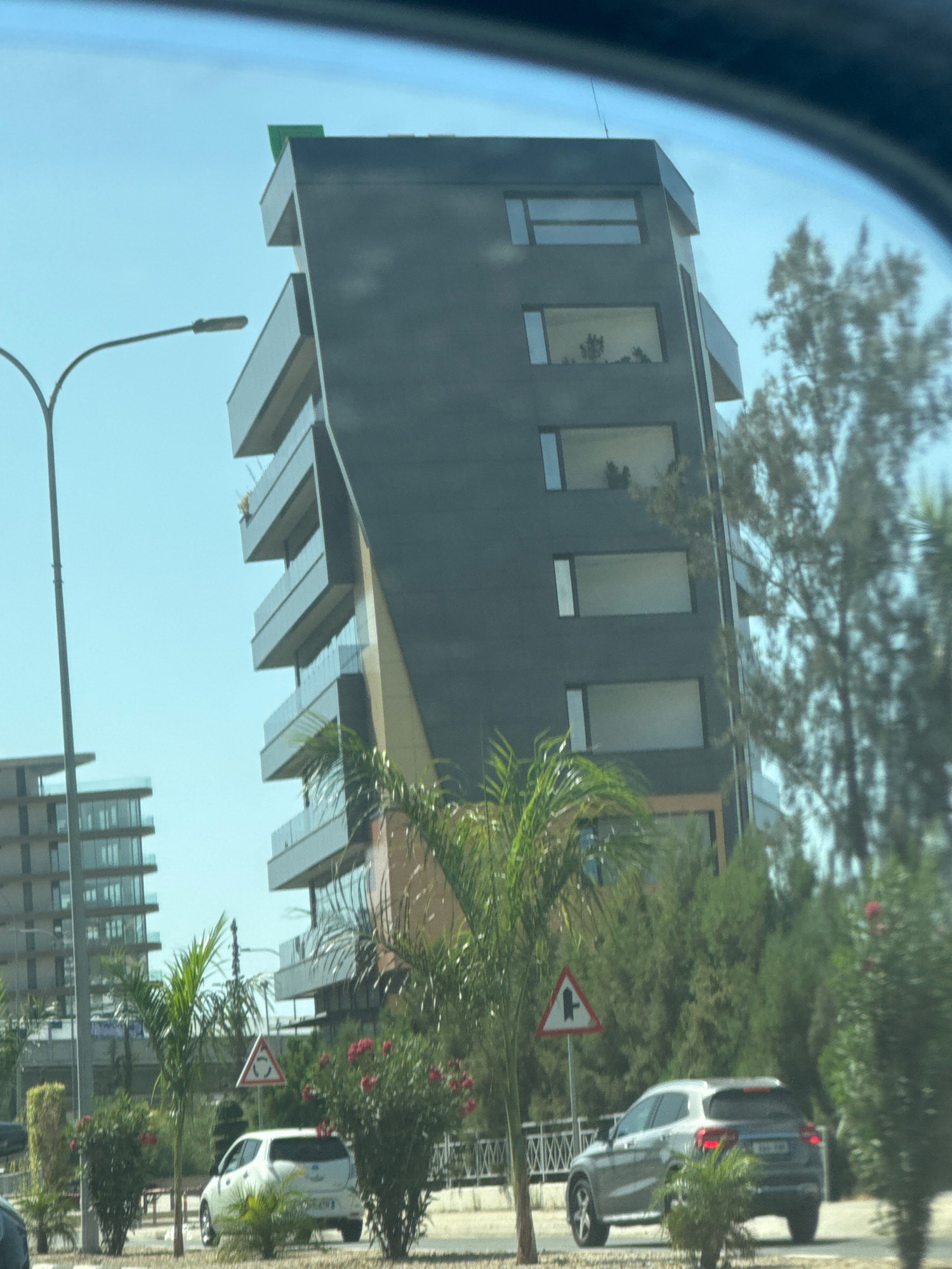 The image shows a modern, multi-story dark gray building with a distinctive angled facade and balconies with glass railings, located in Germasogeia Municipality, Cyprus. A smaller, lighter-colored building with glass windows is visible to the left. The scene includes a streetlamp, lush green trees and palm trees, flowering bushes with red blossoms, and two cars on the road. One car is a white hatchback, and the other is a silver SUV. Traffic signs are visible, including a roundabout sign and a junction sign. The sky is clear and blue, suggesting a sunny day. The image appears to have been taken from a moving vehicle, as evidenced by the slight blur and reflection in the foreground, possibly from a car windshield.