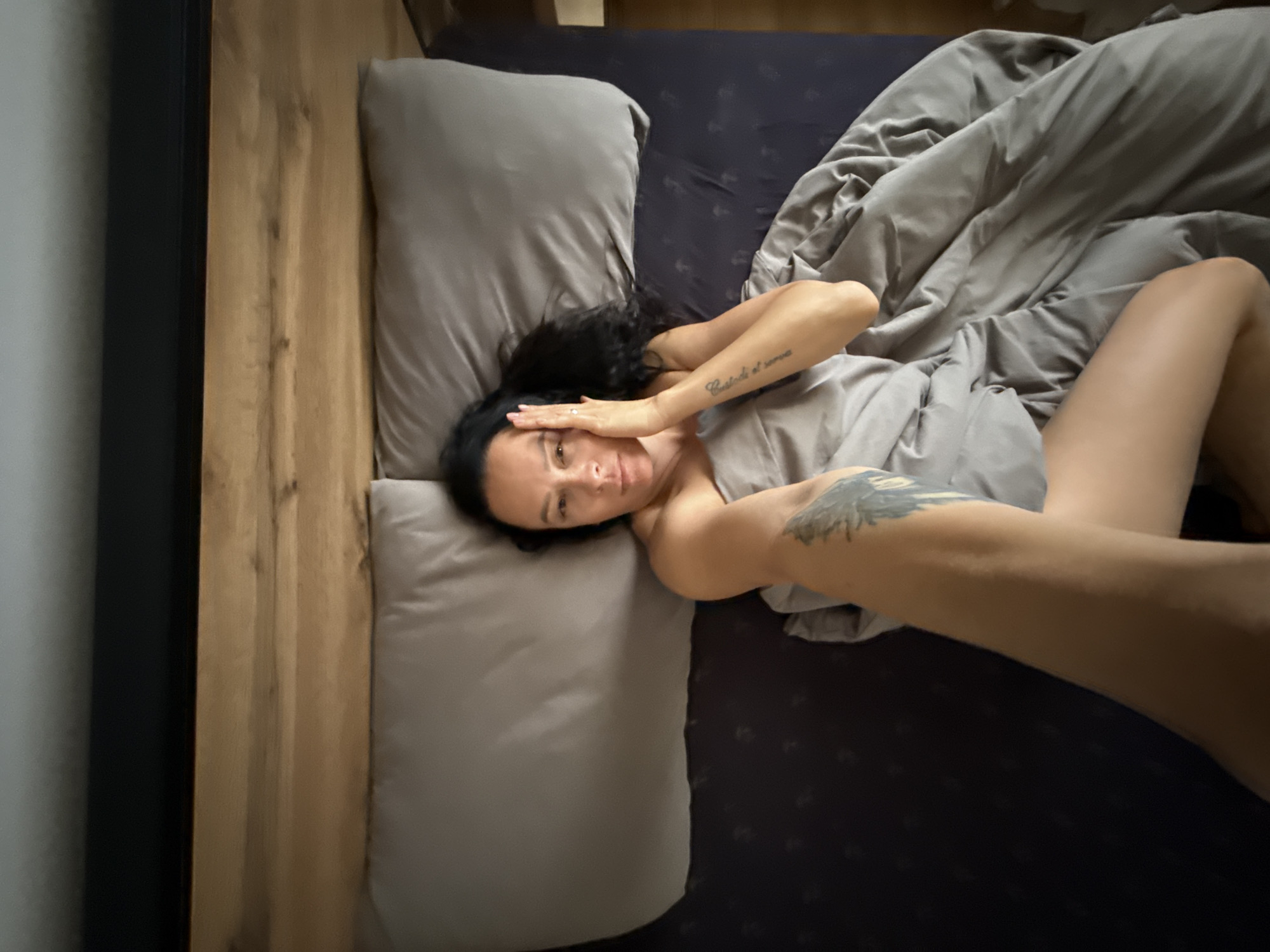A woman is lying on a bed, on her back, with her legs in the air. Her body is partially covered by a grey duvet. She is looking at the camera and has her hands on her face. There is a tattoo on her left arm. The bed is in a room with dark blue sheets and a wooden headboard. The image is taken from a high angle, looking down at the woman. The location is not specified.