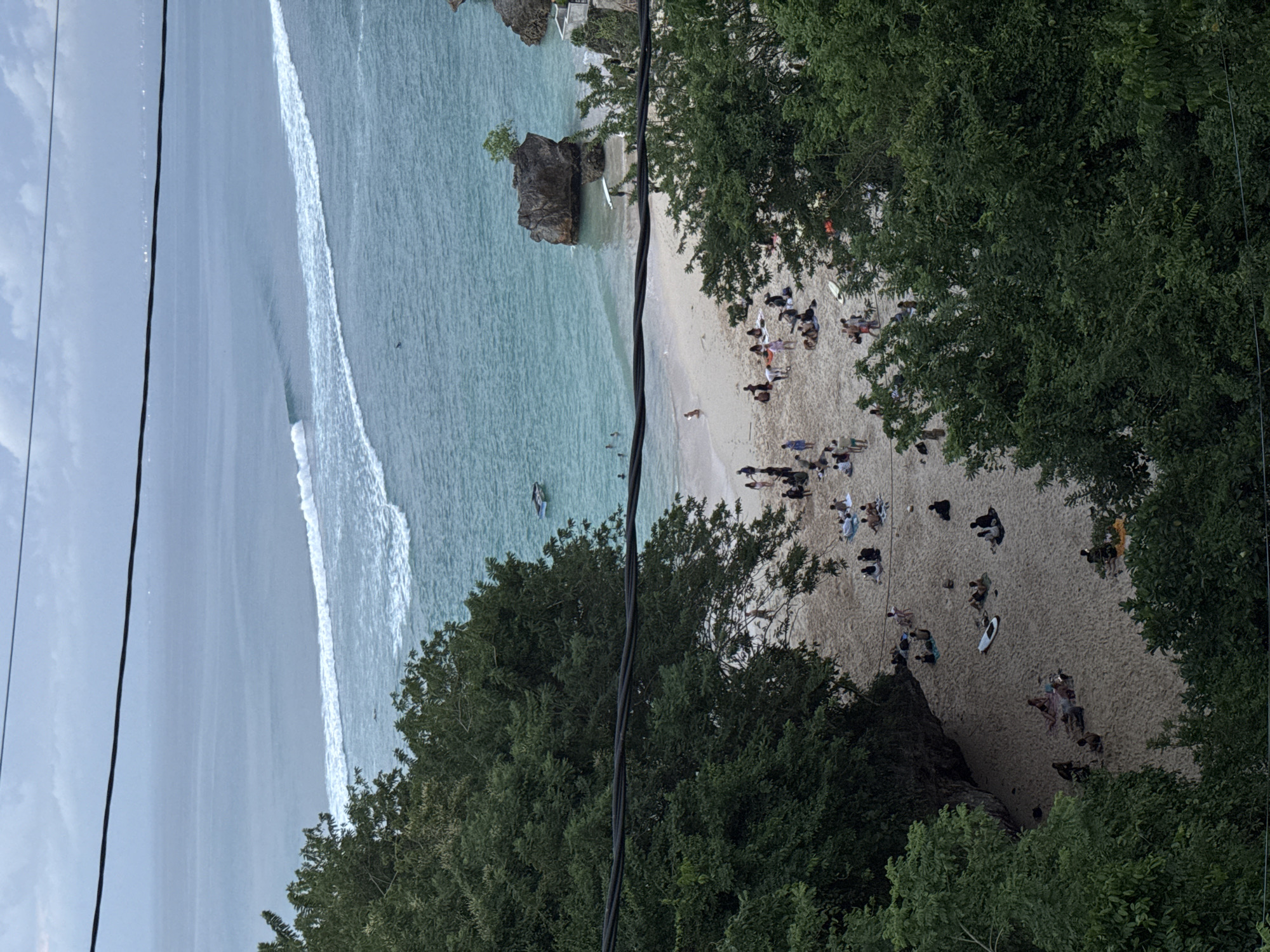 From an elevated vantage point, the image captures a beach scene in Kuta Selatan, Indonesia. A sandy beach is populated with numerous people engaged in various activities like sunbathing, standing, and sitting. In the water, waves break along the shoreline. A prominent rock formation, topped with vegetation, sits at the edge of the beach. Lush green trees frame the beach. Electrical lines are in the foreground.