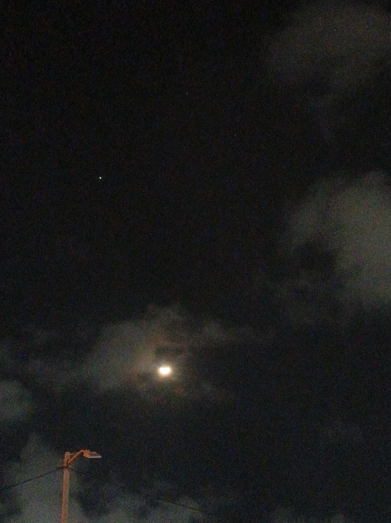 The image captures a nighttime scene over Porlamar, Venezuela, featuring a dark, cloudy sky. A bright moon is visible in the middle-right portion of the sky, partially obscured by translucent clouds but still casting a significant glow that illuminates the surrounding cloud formations. Its visible shape through the clouds suggests it could be in a crescent or gibbous phase.
Two distinct, bright celestial bodies are noticeable against the dark sky: one prominent in the upper left quadrant and another fainter one slightly above and to the left of the moon. These are likely bright stars or planets. In the bottom-left corner of the image, a light brown utility pole with a street light fixture is present, from which power lines extend.
The time of day is clearly night, and the weather is cloudy, yet clear enough to allow some celestial objects to be seen. There are no people visible, nor is there any discernible text within the image. The scene presents a calm, nocturnal landscape blending natural elements with urban infrastructure.