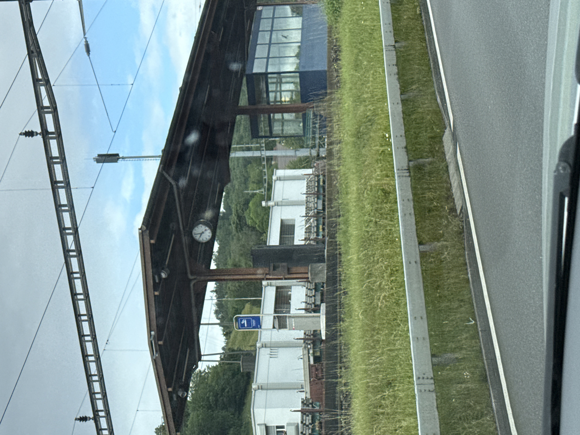 The image captures a sideways view of a railway environment, likely from a moving train or vehicle, looking towards a station platform in Zurzach, Switzerland. When viewed upright, the foreground on the left features intricate overhead catenary lines and support structures for an electric railway.
Central to the image is a train platform covered by a robust, dark-colored canopy. Suspended from the canopy's supports is a circular clock, which indicates the time as approximately 10:10. Beneath the canopy and extending behind it are several white, functional-looking station buildings. A prominent blue rectangular sign, typically used for platform identification, is visible in the midground, displaying a white left-pointing arrow and the number "1".
In the background, lush green hills covered in trees rise behind the station buildings. A modern blue building with large window panes is also discernible, reflecting parts of the cloudy sky. To the right of the platform infrastructure, a paved road or track runs parallel to a neatly maintained grassy verge, separated by a concrete barrier. The sky above is partially overcast with patches of blue visible. No people are apparent in the scene, which appears to depict a typical, quiet regional train station setting.