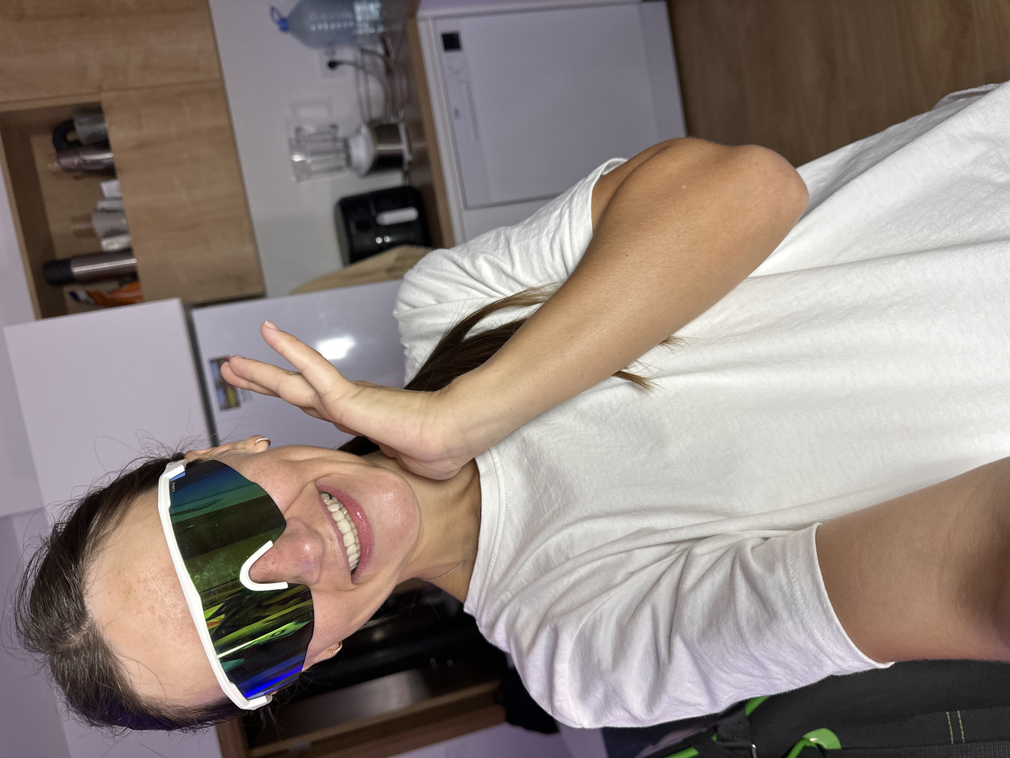 A woman is taking a selfie in a kitchen. She is wearing a white t-shirt and large, wraparound sunglasses with reflective lenses. Her right hand is raised to her face, and she is smiling broadly. Behind her, a kitchen counter is visible with appliances such as a refrigerator, air fryer, blender, and dishwasher. The overall setting appears to be a domestic kitchen, possibly in an apartment or house. The image is taken from a medium shot, focusing on the woman's upper body and face.