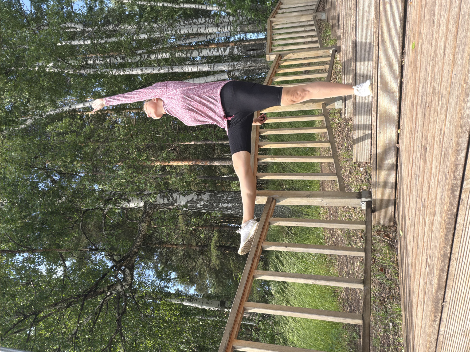 A woman is performing a high leg stretch with her right leg extended horizontally and resting on a wooden railing. She is wearing a pink long-sleeved shirt and black cycling shorts, along with white athletic shoes. Her left arm is raised vertically, reaching upwards, and her head is tilted back, looking towards the sky. The background features a dense forest of birch trees with white bark and green foliage. The woman is positioned on a wooden platform, with wooden stairs leading upwards to her right. The ground is covered with dry leaves and sparse grass. The overall scene suggests an outdoor exercise session in a natural, wooded environment. The location is the area of Chaikovsky, Russia.