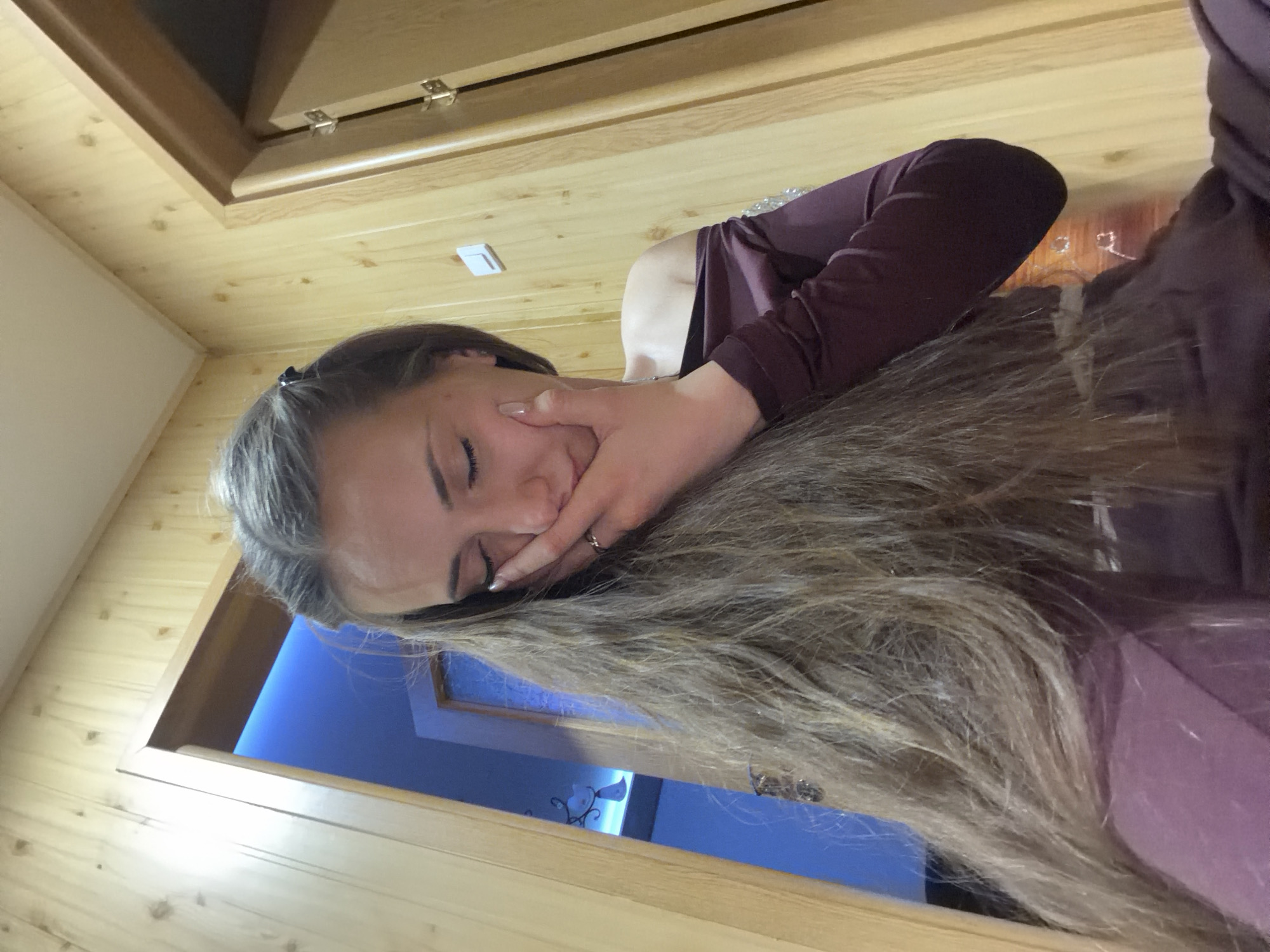 A woman with long blonde hair is posing for a photo. She is wearing a long-sleeved purple shirt and a ring on her left hand. She is holding her hand to her face. The background appears to be the interior of a room with light wood paneling and a blue wall visible through a doorway. The photo appears to have been taken indoors. The location, Buhovo, Bulgaria, is not directly verifiable from the image content.