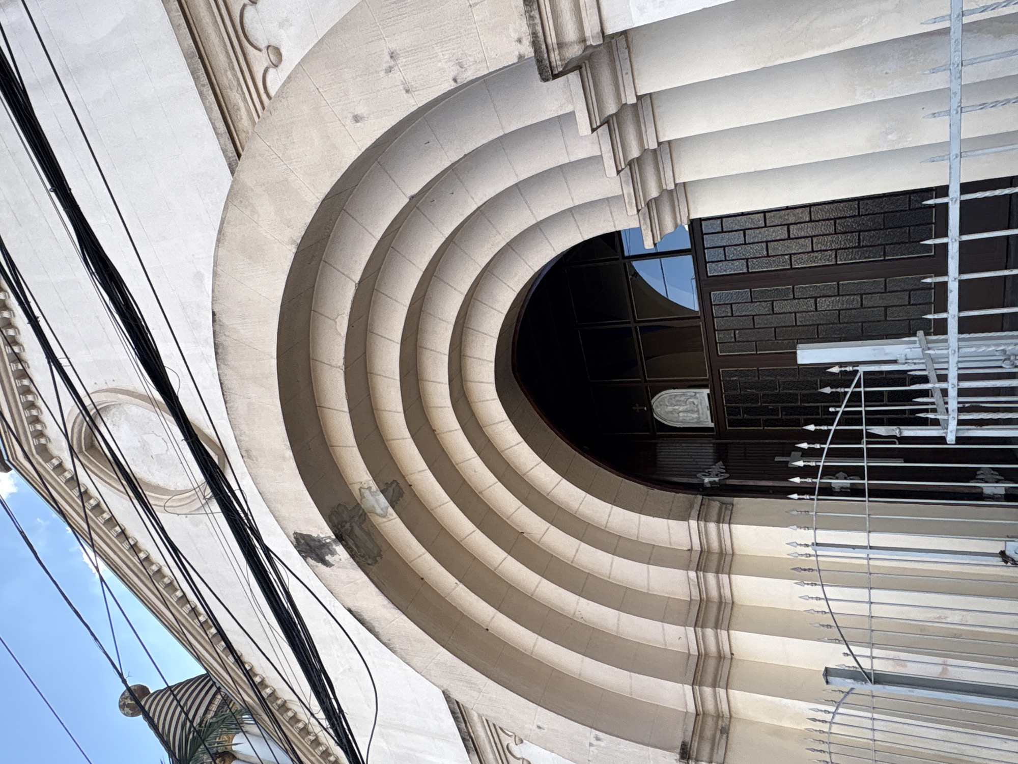 The image shows the facade of a church in the city of São Paulo, Brazil. The church has a prominent arched entrance with concentric stone arches and columns. Above the entrance, the building's facade is adorned with decorative molding and an oval niche. Several black electrical wires are strung across the upper portion of the building, crossing in front of the blue sky. To the left, a conical structure with striped detailing is partially visible, possibly part of the church's architectural design. In front of the entrance, a metal fence with pointed finials encloses the area. The entrance itself features dark, paneled doors with glass inserts, and a small religious relief sculpture is visible above the right door. The overall scene suggests a place of worship with some visible signs of urban infrastructure and wear on the building's exterior.