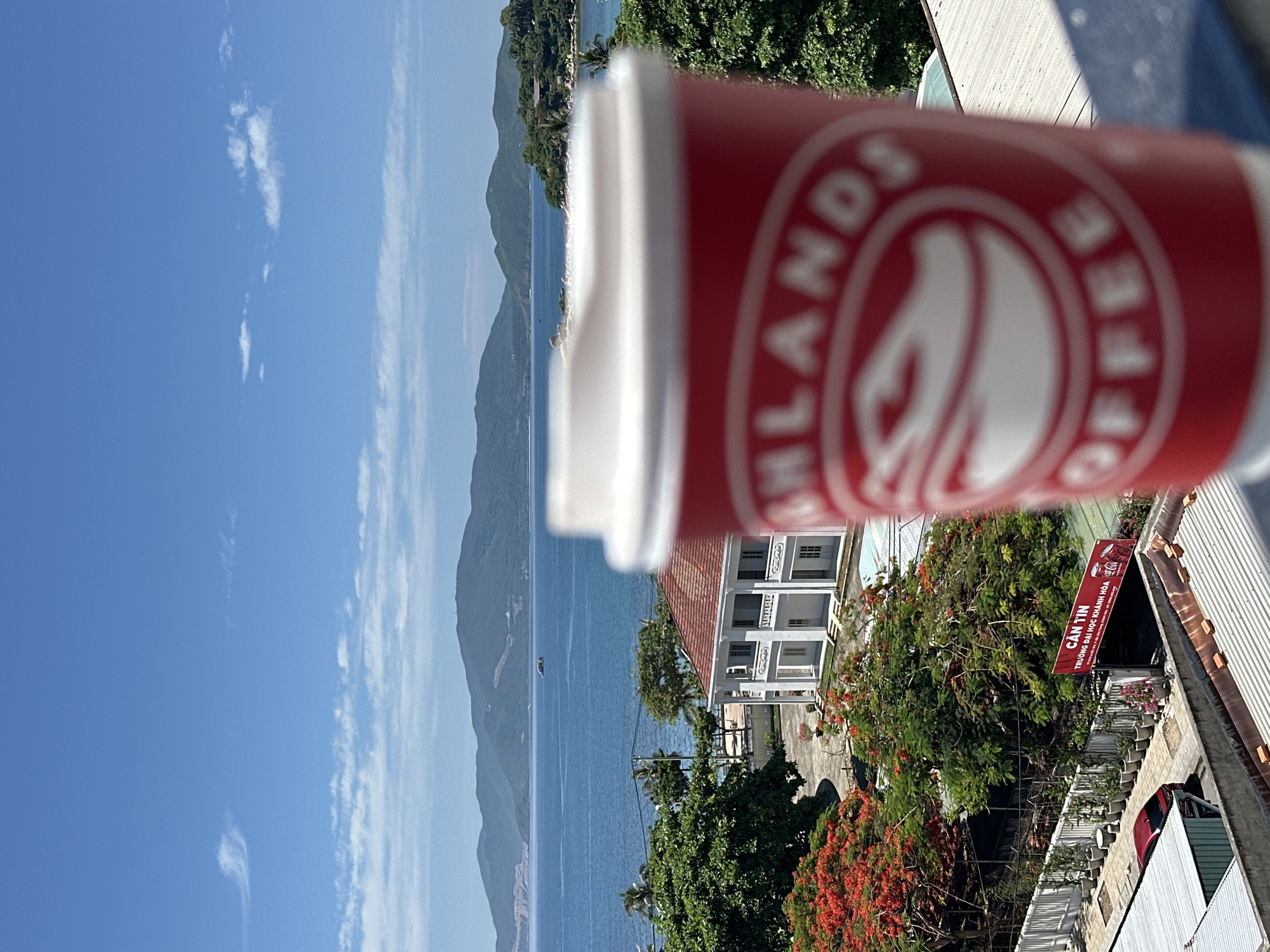 In Nha Trang, Vietnam, a red and white disposable coffee cup from "Highlands Coffee" is in the foreground, out of focus, obscuring the view. The background reveals a scenic coastal landscape under a bright blue sky with scattered white clouds. The ocean stretches out to distant, lush green hills and mountains. A white building with a red-tiled roof is visible amidst dense green foliage, including a tree with vibrant red flowers. A sign in Vietnamese, which reads "CÀN TIN TRƯỜNG ĐẠI HỌC KHÁNH HÒA," indicating a canteen for Khanh Hoa University, is also present. A red car is parked on a paved area. The scene appears to be taken from a higher vantage point, possibly a balcony or rooftop, overlooking a tranquil urban and natural environment.