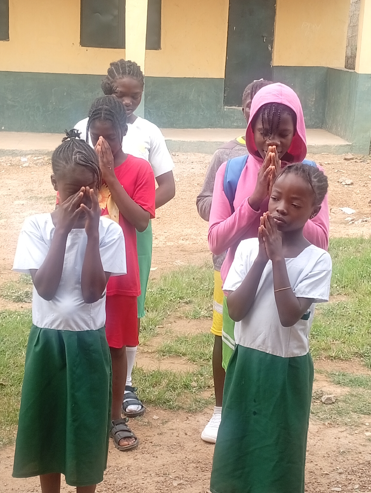 In Narayi, Nigeria, a group of young girls stand in a line outside a building with yellow and green walls. The girls are dressed in school uniforms; some wear white shirts and green skirts, while one wears a red shirt and shorts. They all have their hands clasped in front of their faces as if in prayer or meditation. A girl in a pink hooded jacket stands behind them, also with her hands clasped. The ground is dirt and gravel with patches of grass.