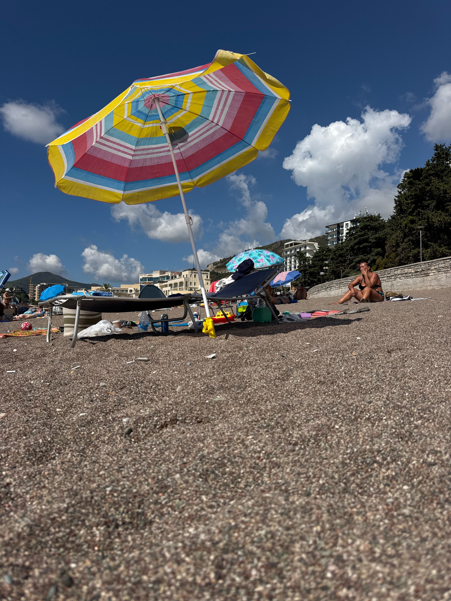 The image depicts a sun-drenched beach scene in Boreti, Montenegro, with a low-angle perspective from the pebble-strewn shore. In the foreground, a large, brightly colored beach umbrella with red, yellow, and blue stripes dominates the upper left portion of the frame, angled towards the sky. Beneath it, several beach chairs and loungers are visible, some occupied and others empty.
Further back, a group of people are scattered across the beach, enjoying the sunny day. A woman in a dark bikini is seated on a towel to the right of the frame, looking towards the left. Other beachgoers are seen relaxing under umbrellas and on loungers.
In the background, buildings, some with white facades and balconies, are nestled against lush green trees that climb a gentle slope. The sky is a vibrant blue, dotted with fluffy white clouds, indicative of a clear summer day. The overall atmosphere is one of leisure and relaxation on a popular coastal destination.