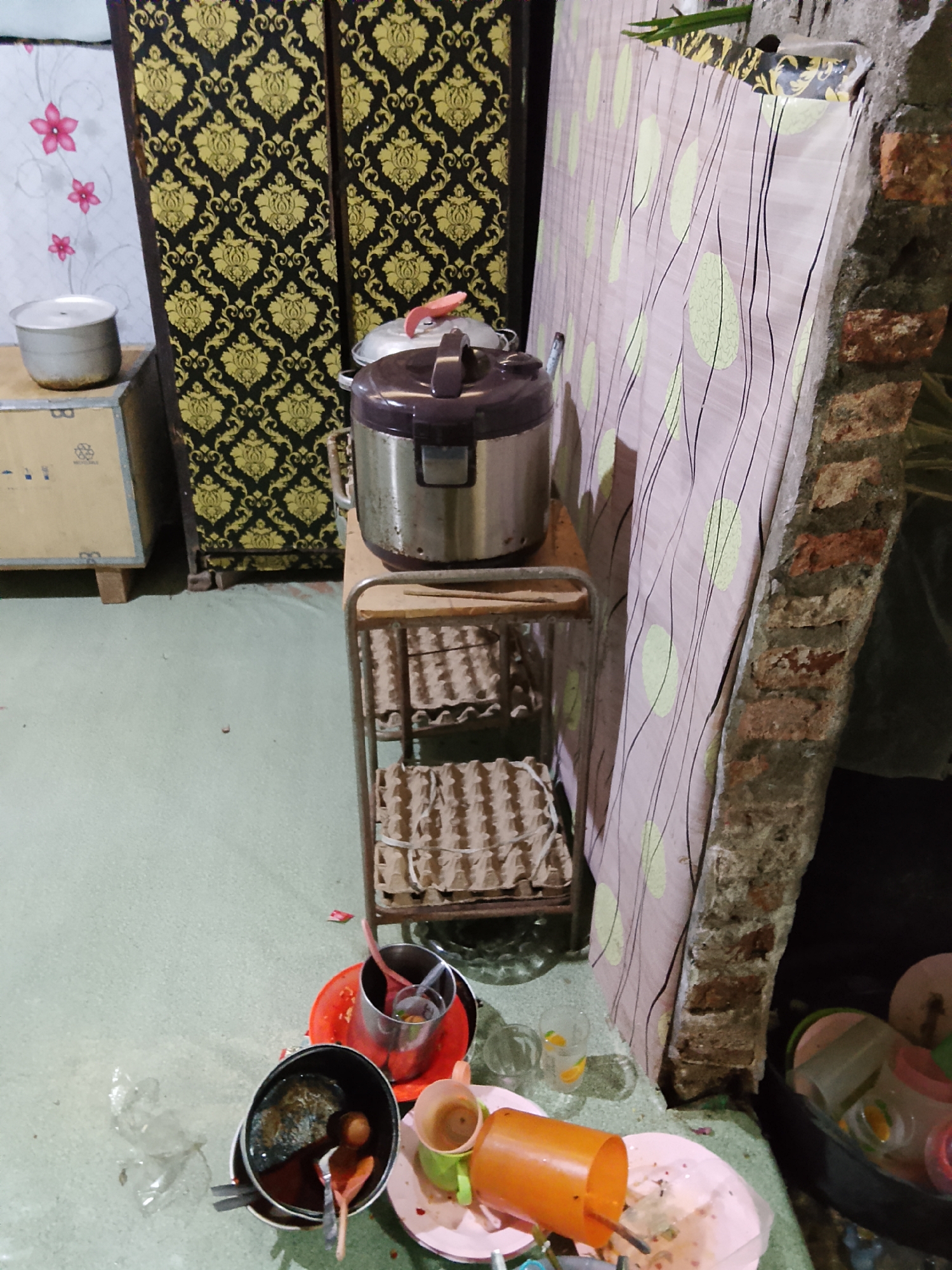 In Merbopuntong, Indonesia, the scene depicts a dimly lit indoor environment, possibly a kitchen or storage area. A large, dark wardrobe with a gold damask pattern stands against a wall adorned with pink floral wallpaper. To its left, a metal pot sits atop a wooden crate. In the center, a metal rack holds multiple tiers of what appear to be egg cartons. A pressure cooker is positioned on the top shelf of the rack. To the right, a large, partially constructed brick wall is covered by a pink sheet with a repeating pattern of light green ovals and black lines. In the foreground, a collection of dirty dishes, cups, and utensils are scattered on the floor, along with a crumpled plastic bag. The floor is a light green color.