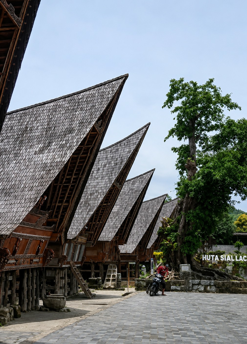The image captures a row of traditional houses with distinct saddle-shaped roofs in Ambarita, Indonesia. The roofs are covered with grey, overlapping shingles. The houses are elevated on stilts and feature ornate wooden carvings on their facades. A paved walkway runs in front of the houses. In the foreground, a person wearing a red shirt and dark pants is standing next to a motorcycle. To the right of the houses, a large tree with dense green foliage stands prominently. In the background, stone structures are visible, with a sign that reads "HUTA SIAL LAC." The sky is clear and blue.
