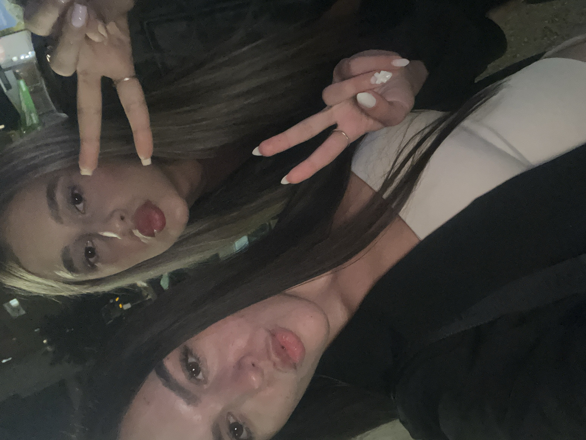 A tilted, low-light selfie captures two young women posing playfully, likely at night in an outdoor setting in Sofia, Bulgaria. The image is taken from a slightly elevated angle.
The woman at the top of the frame has blonde hair, parted to the side, and is looking directly at the camera with a slight smile, sticking her tongue out. She wears a dark top. Her right hand is raised, making a peace sign (V-shape) with fingers spread, revealing light-colored nail polish. A ring is visible on her ring finger.
The woman at the bottom of the frame has long, dark hair. She is also looking at the camera, puckering her lips. She wears a white top underneath a dark jacket. Her left hand is raised, mirroring the peace sign, showcasing white nail polish, with a distinct white flower design on one nail. She also wears a ring on her ring finger.
The background is dark and blurred, with faint artificial lights and what appears to be bottles or glasses, suggesting a social venue such as a bar or restaurant patio. Both women exhibit a casual and friendly demeanor, consistent with a shared moment captured in a selfie. There is no visible text in the image.
