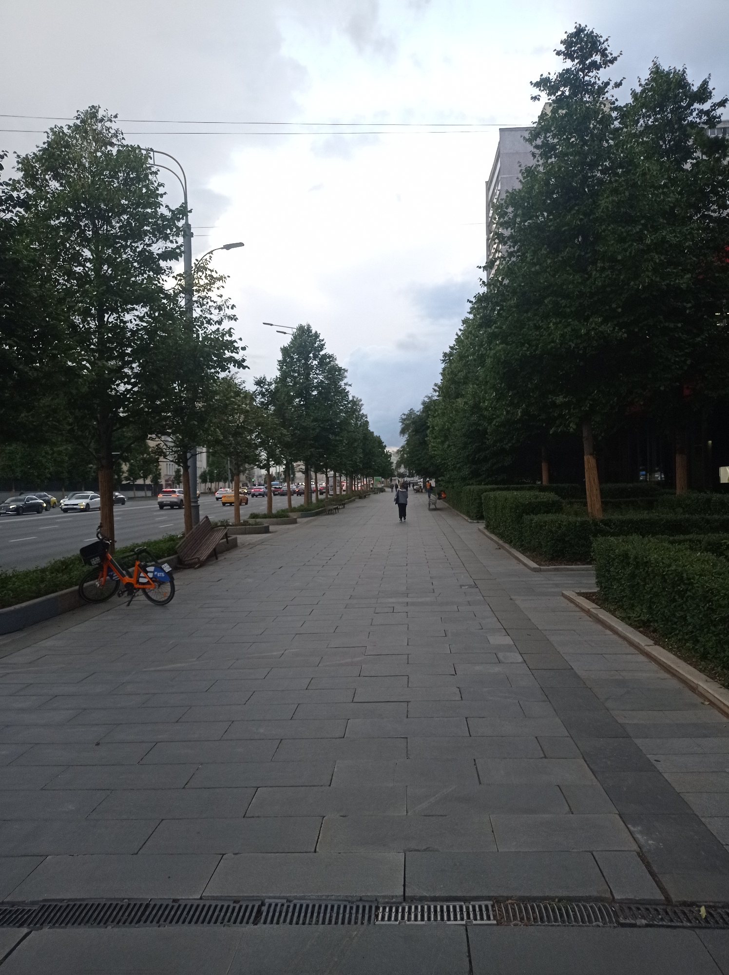 A wide, paved pedestrian walkway stretches into the distance along a city street lined with trees and buildings. The walkway is constructed of large, rectangular grey paving stones. A metal grate drain runs along the edge of the walkway near the foreground. To the left of the walkway is a street with multiple lanes of traffic, including cars and a yellow taxi. A few parked cars are visible on the side of the street. An orange bicycle, possibly a shared rental bike, is parked on the walkway near the left edge, leaning against a small bush. A wooden park bench is positioned behind the bicycle. Further down the walkway, a lone person in dark clothing with a backpack is walking away from the camera. The right side of the walkway is bordered by neatly trimmed green hedges. In the background, tall trees with dense green foliage line the street, and modern buildings with windows are visible behind them. The sky is overcast with grey clouds. The scene suggests an urban environment in Moscow, Russia, likely on a cloudy day.