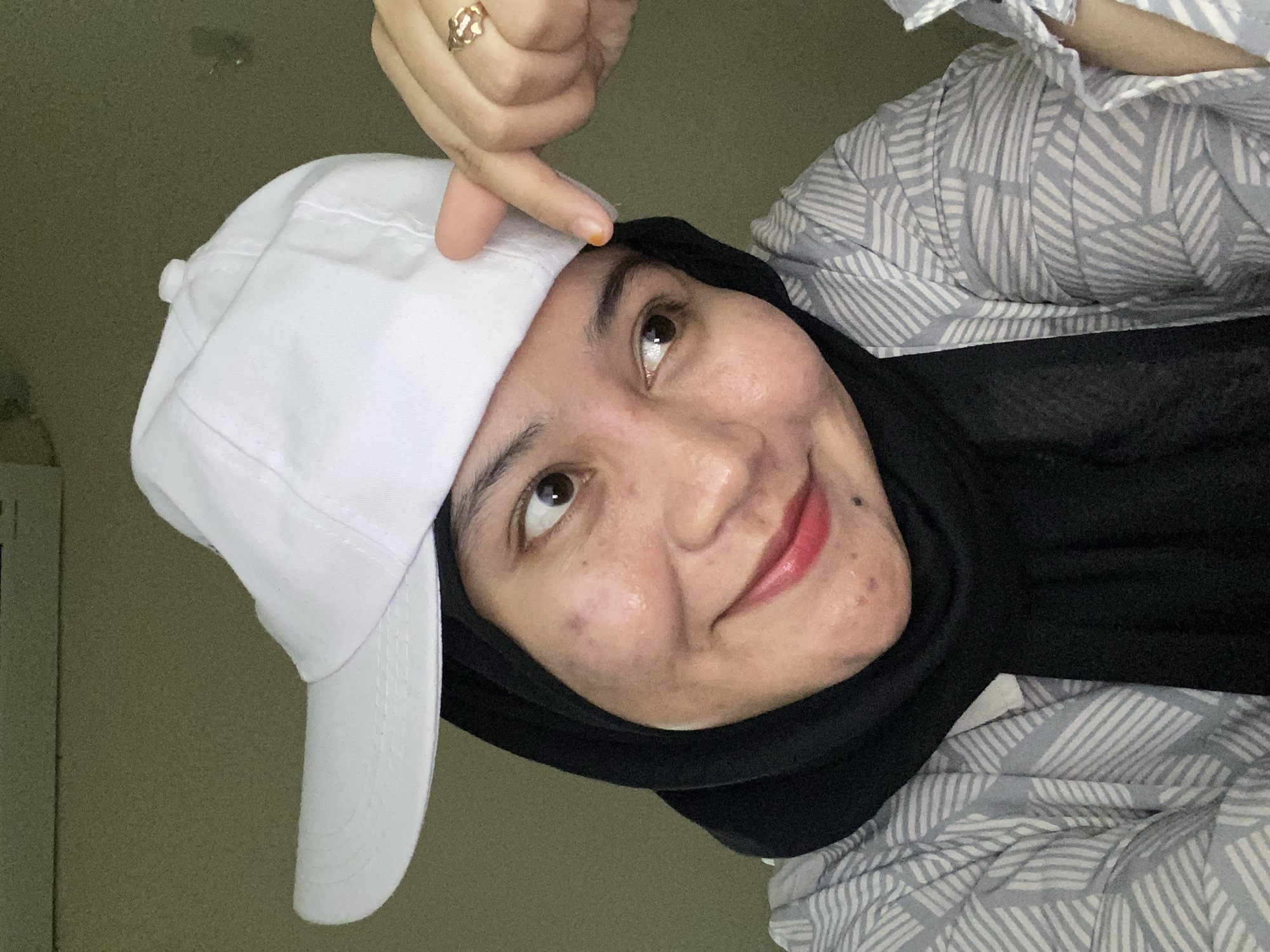 A woman wearing a black hijab, a white baseball cap, and a patterned shirt poses for a selfie. Her left hand is raised, with her index finger pointing at the brim of her cap. She is wearing a gold-colored ring on her right hand. The background appears to be an indoor setting with a plain, neutral-colored wall. The lighting suggests it is likely daytime. The image was captured in Banjarbaru, Indonesia.