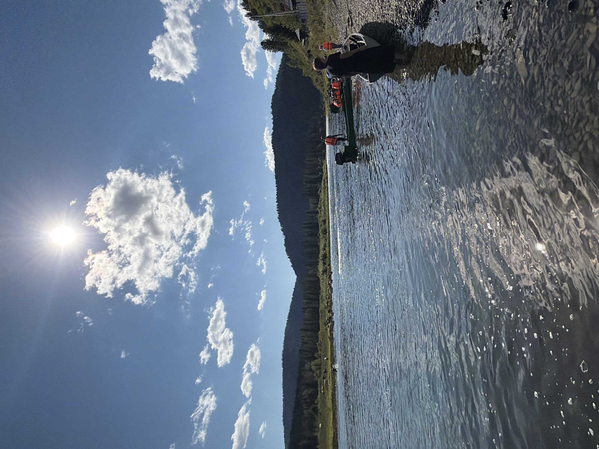 The image depicts a sunny day on the bank of the Bolshoy Izyuk River in Ust-Kabirza, Russia. The river is wide and calm, with sunlight glinting off its surface. The foreground is a rocky shoreline. In the middle ground, a group of people are gathered around two boats. One boat, a green motorboat, is in the water and has several people wearing orange life vests inside. Another boat, a canoe, is partially pulled onto the shore. A person in a black dress is standing next to the canoe, facing away from the camera. In the background, rolling hills covered in dark green pine forests rise against a bright blue sky dotted with fluffy white clouds. The sun is high in the sky, casting a bright glare.