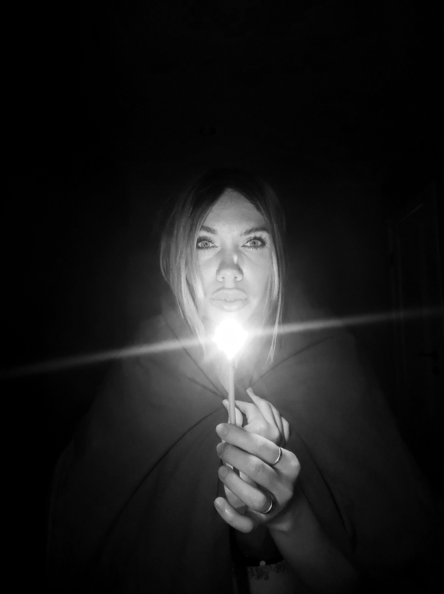 A black and white photograph shows a person holding a lit candle in front of their face. The person is wearing a dark garment, possibly a cloak or robe. The light from the candle illuminates the person's face, particularly their eyes and lips. The background is dark. The photograph does not reveal details related to a specific location, such as Votkinsk, Russia.