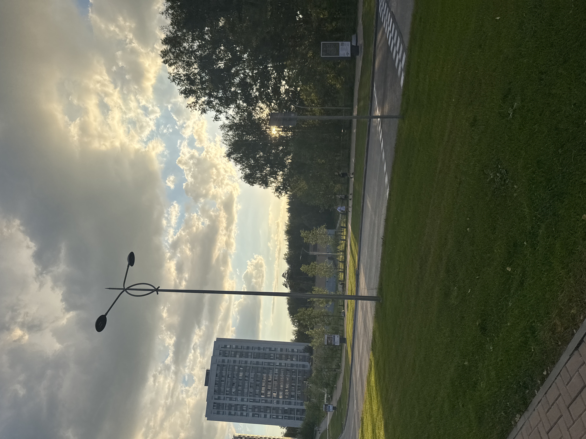 This is a vertical, full-shot photograph of a street scene in Koltsovo, Russia, likely taken during the late afternoon or early evening due to the warm, angled sunlight. The sky is filled with dramatic, fluffy clouds, illuminated by the sun which is partially obscured by trees, creating a luminous effect.
The foreground on the right is dominated by a lush green lawn bordered by a brick pathway. To the left of the lawn is a paved road with white dashed lines and a pedestrian crossing marked by black and white stripes. A modern streetlamp with three oval lights stands prominently in the middle left of the frame. Further back, a tall, multi-story residential building with numerous windows is visible. Beyond the road and the building, a dense line of trees forms the background, with some smaller trees and bushes lining the road.
There are a few people visible on the pedestrian path near the road, but they are small and not the focus of the image. The overall mood is serene and perhaps a little dramatic due to the sky's appearance.
There is some text visible on a signpost near the trees, but it is too small to be legible.