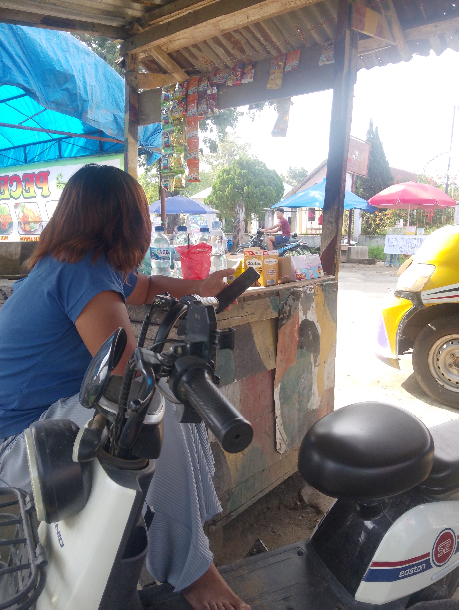 A woman in a blue t-shirt and a grey striped skirt sits on a white and black scooter, facing away from the camera. She is holding a black smartphone in her right hand. The scooter is parked in front of a roadside stall with a corrugated metal roof supported by wooden posts. Various snacks are displayed hanging from the roof. Behind the stall, several bottles of water and red plastic bags are visible. In the background, a young man on a blue motorcycle rides past. Further back, a yellow vehicle with an umbrella in front of it is visible. The scene is set in a street in Serbelawan, Indonesia, with trees and buildings in the distance. The lighting suggests it is daytime, with bright sunlight.
