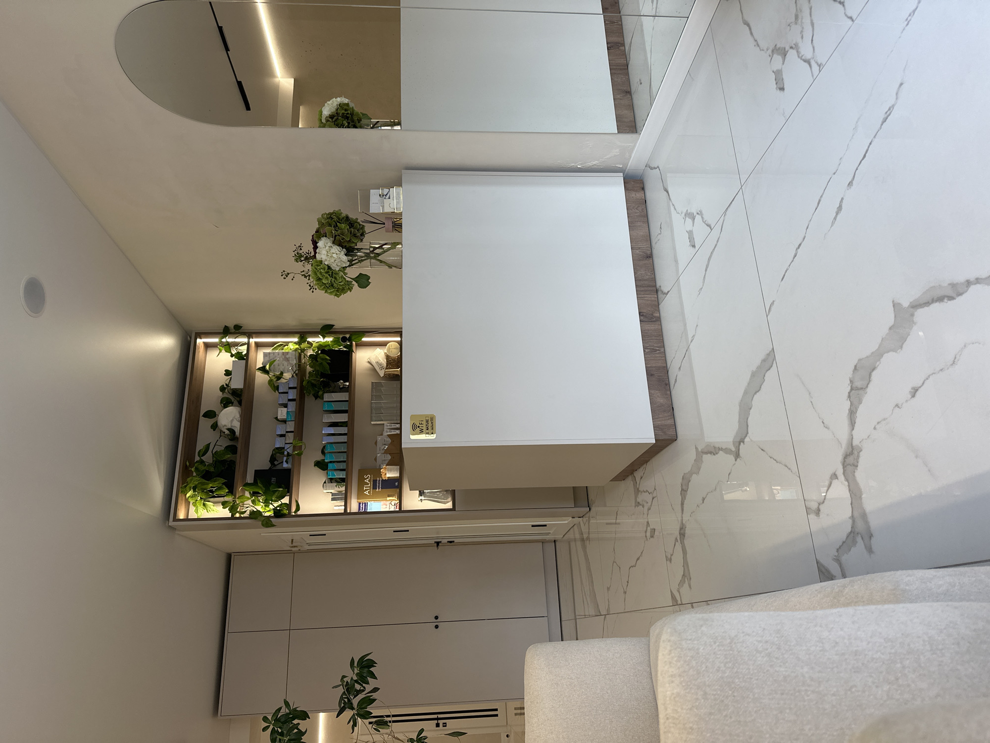 The image shows the interior of a modern, well-lit reception area, likely a spa or salon, with a clean, minimalist aesthetic. A white reception desk with a wooden base is positioned centrally. Behind it, a shelf unit displays various skincare products and plants. To the left, a closet with white doors is visible. The floor is covered in large, glossy white tiles with grey marbling. To the right, a large arched mirror reflects the room. In the foreground, the armrest of a plush, light-colored armchair is visible. The environment appears to be in Ternopil, Ukraine, suggesting a commercial establishment within the city. There are no people present in the image.