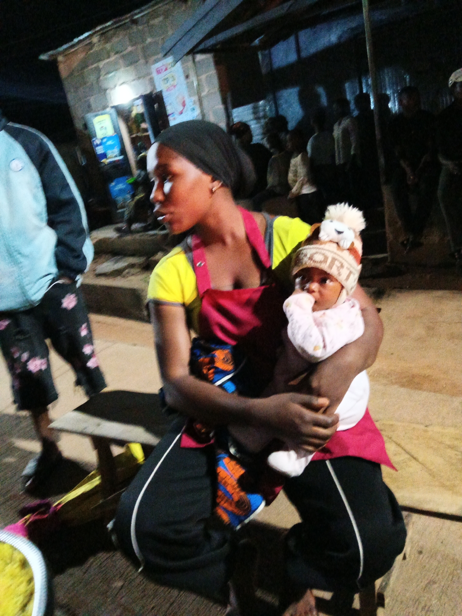 In Bukuru, Nigeria, a woman in a yellow shirt and red apron holds a baby wearing a pink onesie and a striped beanie. The woman is seated on a bench. In the background, a person in a light blue jacket and black shorts with pink floral patterns stands. The environment appears to be an outdoor market or roadside stall, with a brick building and other individuals visible in the background, suggesting a bustling urban scene at night.