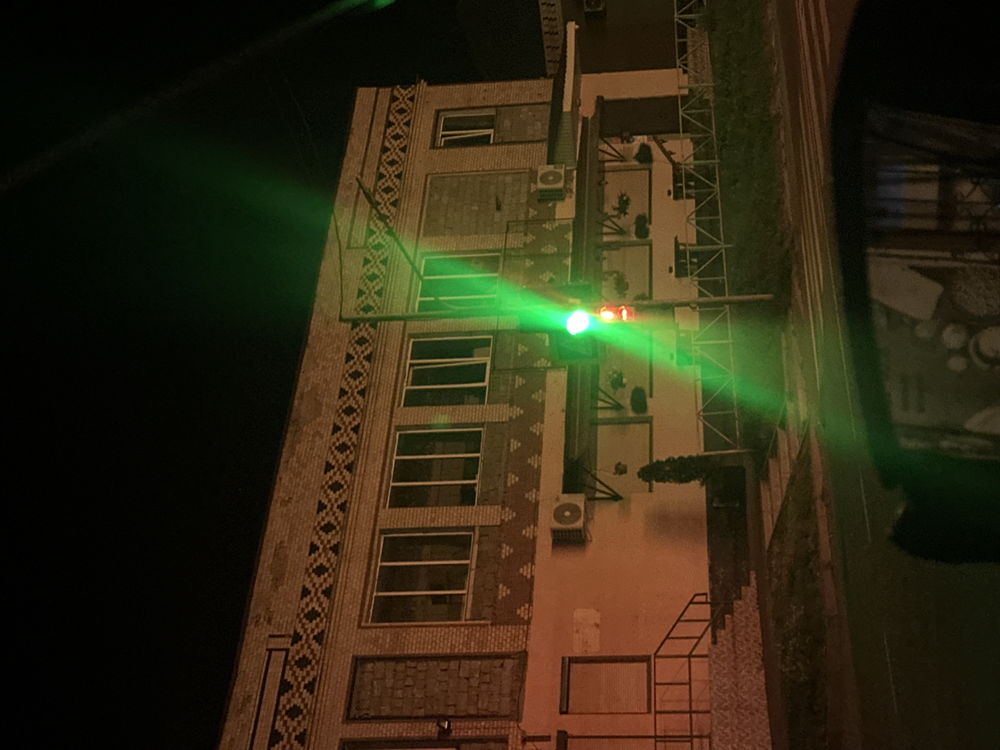 The image captures a nighttime scene of a building and a traffic light, with a bright green light from the traffic signal dominating the foreground and casting a vibrant glow. The building appears to be a two-story structure made of brick with decorative elements along the facade. There are several windows visible, some of which are boarded up or covered. Air conditioning units are mounted on the exterior walls. A set of stairs leads up to an entrance on the left side of the building. The street in front of the building has a pedestrian crossing marked by white stripes. The overall environment suggests an urban setting, possibly in Shatava, Ukraine, given the provided location information. The photograph is taken from a low angle, possibly from within a vehicle, as a reflection is visible in what appears to be a car's side mirror in the bottom portion of the image.