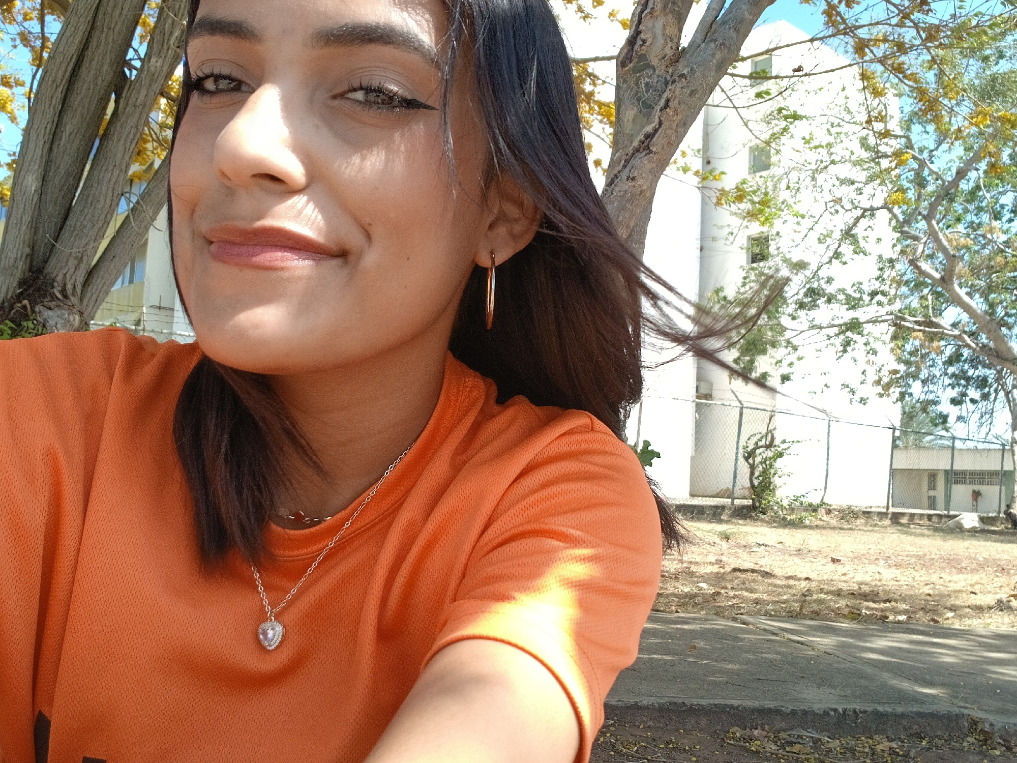 A woman with dark hair is taking a selfie outdoors. She is wearing an orange t-shirt and a silver necklace with a heart-shaped pendant. She has a gold hoop earring in her left ear. Behind her, there is a tree with yellow flowers and a white building. A chain-link fence and a grassy area are also visible. The location is Altagracia, Venezuela.