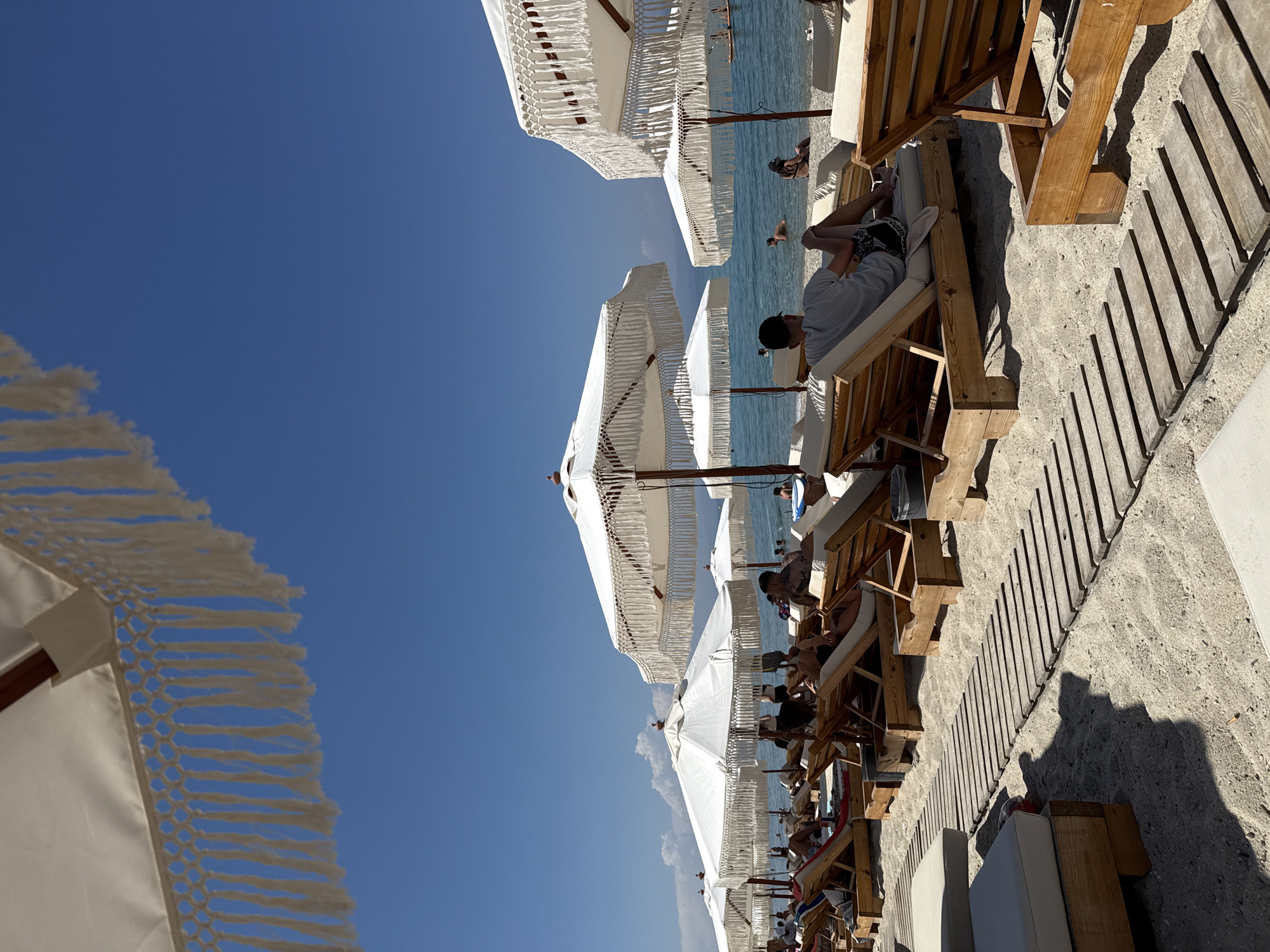 The image depicts a sunny day at a beach resort in Pallini, Greece. The scene is oriented vertically, with the sky taking up a significant portion of the left side of the frame. A clear blue sky dominates the upper left, with a few wispy white clouds visible in the lower left quadrant.
The foreground and midground are filled with rows of wooden lounge chairs and white beach umbrellas. The umbrellas are distinct, featuring fringe detailing along their edges. Several people are visible relaxing on the lounge chairs, some appearing to be reading or simply enjoying the sun. The chairs are arranged on a sandy beach, with a wooden walkway running parallel to them in the right half of the image. The sea is visible in the background, appearing calm and turquoise.
The time of day appears to be midday or early afternoon, given the bright sunlight and the shadows cast by the umbrellas and chairs. The weather is clearly sunny and warm, ideal for a beach outing. The overall atmosphere conveyed is one of leisure and relaxation. No visible text is present in the image.