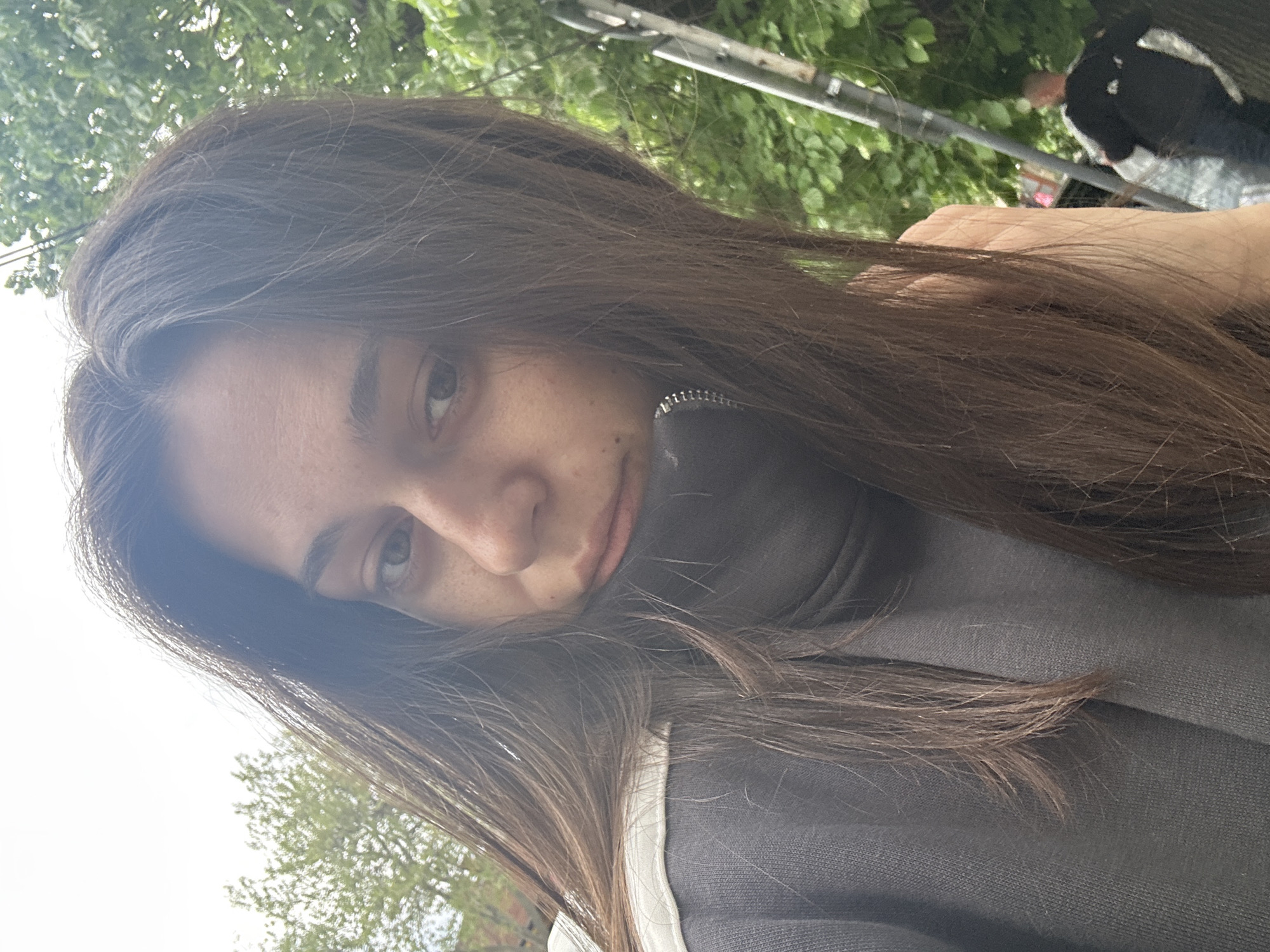 A close-up, slightly tilted portrait shows a young woman outdoors in Veliko Tarnovo, Bulgaria, during the daytime. Her head is angled, with her long, dark brown hair falling across her shoulders and chest. Her left eye, eyebrow, and the bridge of her nose, accented with light freckles, are clearly visible. Her lips are slightly parted, forming a soft, neutral expression.
She is wearing a dark gray or charcoal-colored zippered jacket or sweatshirt, with a hint of white trim or an undershirt visible near her right shoulder. Her hair has subtle lighter streaks or highlights, catching the natural light.
The background is bright and green, consisting primarily of blurred foliage from trees and bushes, indicating a natural or park-like setting. Patches of bright sky are visible through the leaves, suggesting a clear day. In the upper right corner, a blurred horizontal metal structure, possibly part of a fence or utility pole, cuts across the greenery. Behind it, a very blurry figure wearing a black and white garment is faintly discernible amidst the foliage. No text is visible in the image.