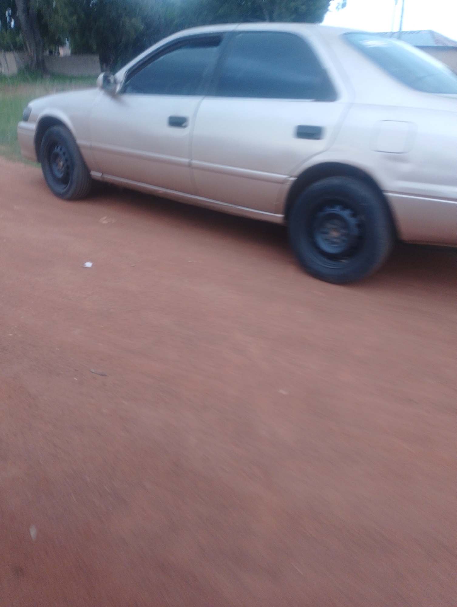 A light-colored sedan is parked on a dirt road in Gura Top, Nigeria. The car is facing right and the image is taken from a low angle, looking towards the passenger side of the vehicle. The front passenger wheel and the rear passenger wheel are visible. The car is clean and appears to be in good condition. The road is made of red dirt and is covered in a thin layer of dust. There are trees and bushes in the background, and a few buildings are visible in the distance. The sky is cloudy.
