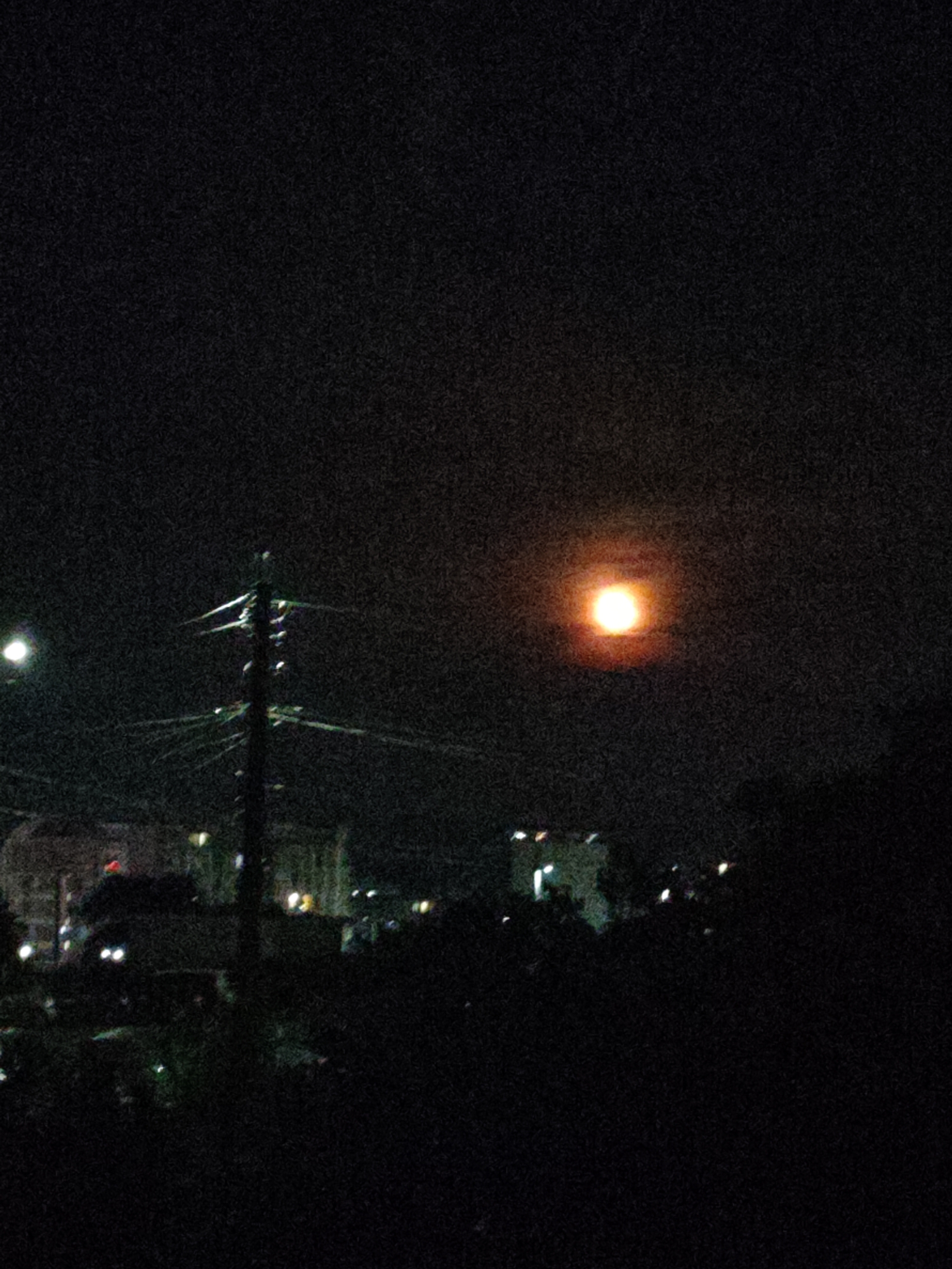 The image captures a dark, grainy night scene in what appears to be an urban or suburban area, likely in Burgas, Bulgaria. The dominant feature in the upper right quadrant is a bright, reddish-orange orb, possibly the moon obscured by atmospheric conditions or a very strong artificial light source, surrounded by a soft halo against the heavily textured black sky.
In the mid-ground, slightly to the left of the center, a dark utility pole stands prominently, adorned with numerous horizontal and diagonal wires. To its immediate left, a distinct, bright white circular light, likely a streetlamp, casts a soft glow. Below and behind these elements, the silhouettes of buildings are visible, dotted with various small, bright lights that suggest windows or other urban illumination. The foreground and lower parts of the mid-ground are consumed by dark, indistinct shapes, likely trees or bushes, heavily shadowed against the minimal ambient light.
The entire photograph exhibits significant digital noise or grain, characteristic of low-light photography, which renders details blurry and textures uneven. No specific activity is depicted, making it a static landscape shot. There is no visible text in the image. The time of day is clearly night, and the scene implies a developed residential or commercial area.
