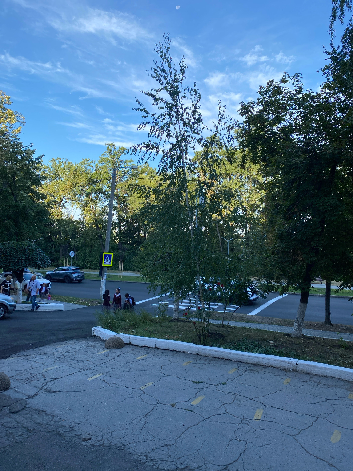 The image depicts a street scene in Chișinău, Moldova, on a clear day with a bright blue sky and wispy clouds. In the foreground, a cracked asphalt surface marked with yellow lines suggests a parking area or a designated zone. A white curb separates this area from a sidewalk and a road.
Several people are visible. On the left side, a group of individuals, some appearing to be adults and a child, are walking. Further down the street, a few school-aged children in uniforms are walking towards the right side of the frame. They appear to be students, possibly heading to or from school.
A dark-colored car is parked on the left side of the road, and another car is visible in the background crossing a pedestrian crossing. Traffic signs are present, including a speed limit sign (30) and a yellow pedestrian crossing sign. The environment is characterized by lush green trees lining the street and a light pole situated near the road. The overall impression is a typical urban street setting in Chișinău, likely during the daytime.
