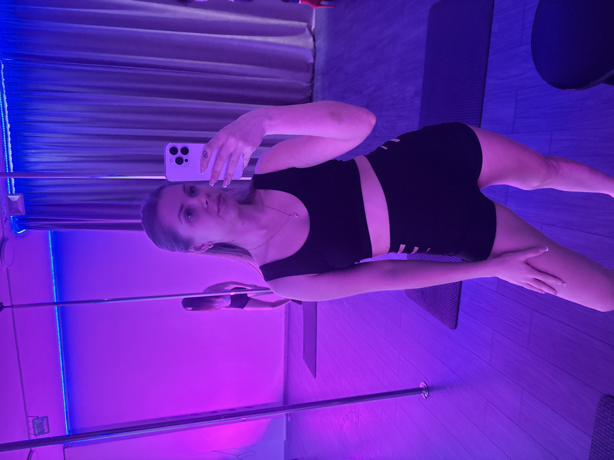 A young woman is taking a selfie in a room lit with blue and purple lights. She is wearing a black sports bra and shorts. A silver pole extends from the floor to the ceiling near the left side of the frame. A mirror is visible behind her. A dark mat lies on the floor beside her. The location is potentially a dance or fitness studio. The city of Batumi, Georgia, cannot be confirmed based on visual evidence alone.