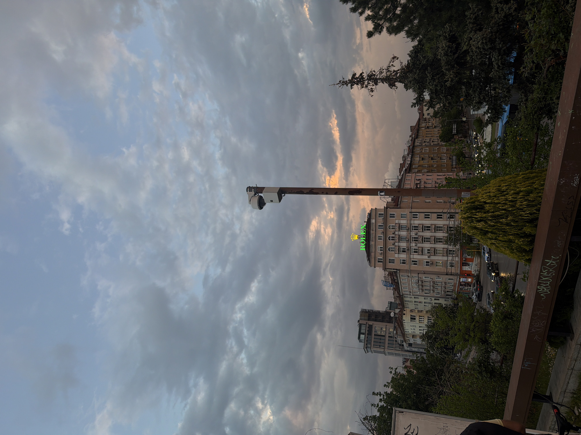 The image shows a street scene in Sofia, Bulgaria. The sky is filled with gray clouds with some hints of sunset on the horizon. In the foreground, there is a security camera mounted on a pole. In the midground, there are buildings, including one with a green Rolex sign on the roof. There are trees and some vegetation in the foreground. The photo seems to have been taken from a high vantage point, like a bridge. There are cars on the street, and the buildings are typical of the city's architecture.