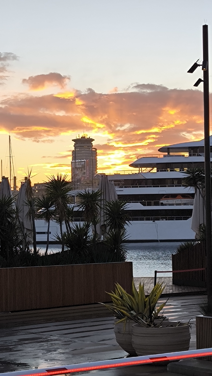 A picturesque harbor scene unfolds with a brilliant sunset illuminating the sky. A tall building, partially obscured by palm trees and yacht masts, stands adjacent to a multi-tiered, luxurious white yacht. The foreground reveals potted plants on a wet, tiled walkway marked by a red LED strip. A security pole with lights is present on the right, completing the serene yet sophisticated ambiance.