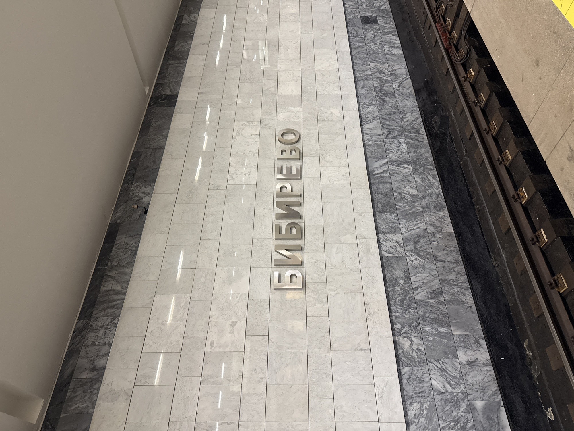 This image shows a section of the Bibirevo metro station in Moscow, Russia. The station name "БИБИРЕВО" is displayed vertically in metal lettering on the light gray, tiled floor. To the right of the tiled area is a section with gray marble-like flooring, and adjacent to that are the metro tracks. The wall on the left is a light gray. The image is taken from an elevated perspective, looking down at an angle.