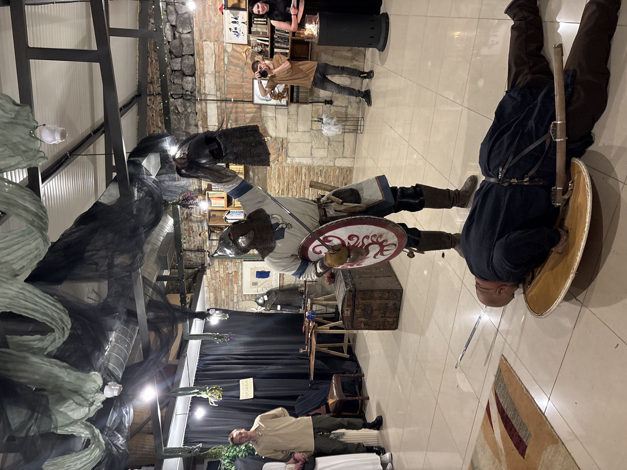 The image captures a scene in what appears to be an indoor event location, possibly in Varna, Bulgaria, given the prompt. The setting has stone and brick wall features with decorative elements such as hanging lights and drapery.
There are several people present. In the foreground, a person dressed in medieval armor is prominently displayed. Nearby, a person appears to be lying face-down on the floor, holding a shield.
In the background, other individuals are seen, one taking a photo with a camera, and others standing by what looks like a fireplace or decorative stove.
Objects within the scene include a sword, a wooden chest, and various decorative items that suggest a themed event or historical reenactment.