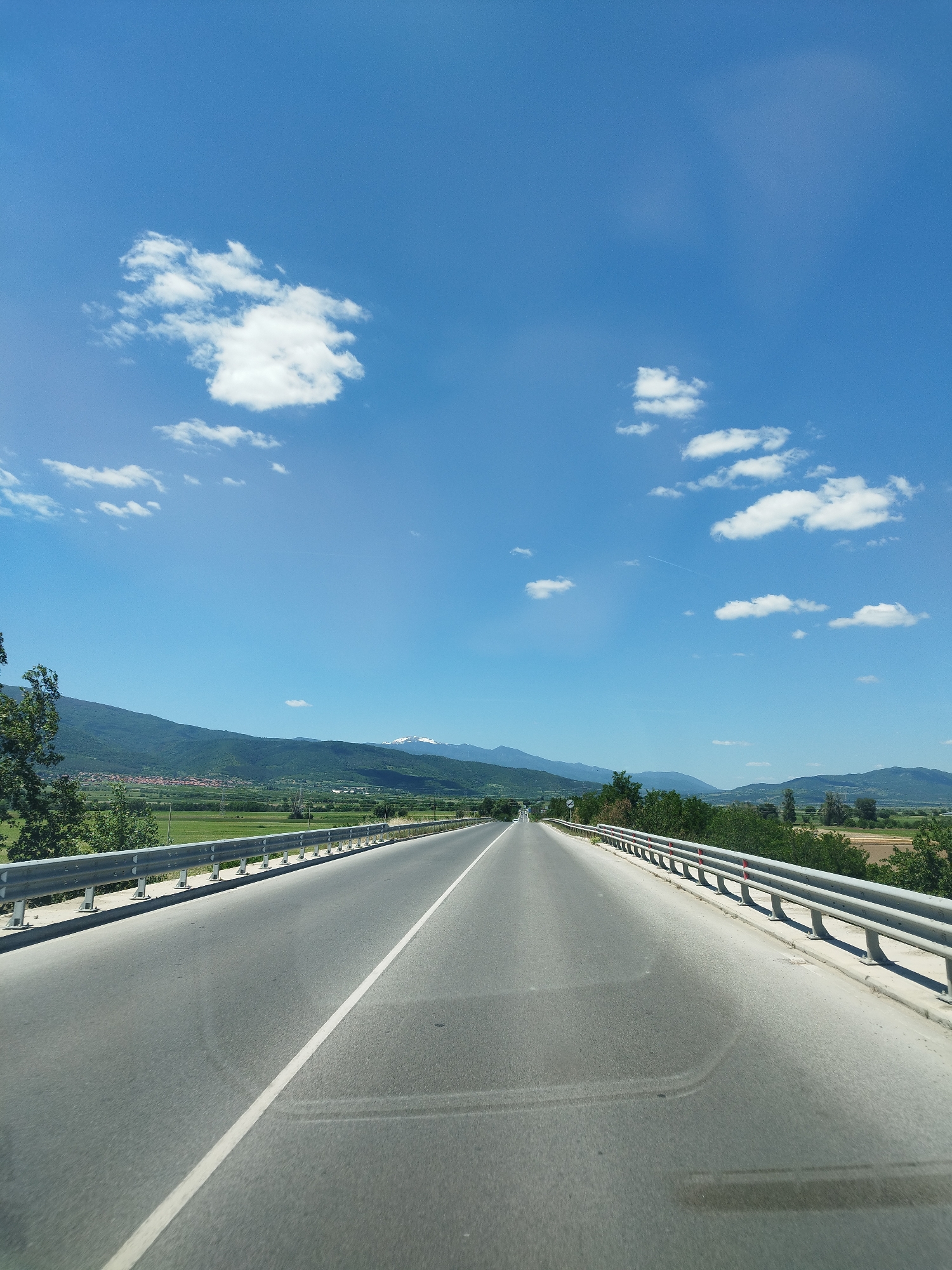 The image shows a straight, asphalt road stretching into the distance under a clear blue sky with scattered white clouds. A white line divides the road. Metal guardrails line both sides of the road. Beyond the guardrails are green fields and trees, with mountains visible in the background. A small town or village is nestled against the mountains. The image appears to be taken from inside a vehicle, looking forward. The location is near Septemvri, Bulgaria, based on the landscape and architectural styles.