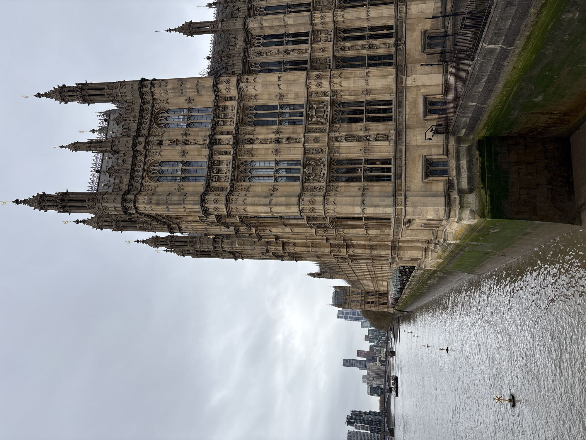 The image depicts the Houses of Parliament in London, United Kingdom, seen from across the River Thames. The architectural details of the building are visible, including its Gothic Revival style, windows, and spires. The river is in the foreground, and other buildings can be seen across the river. The sky is overcast.