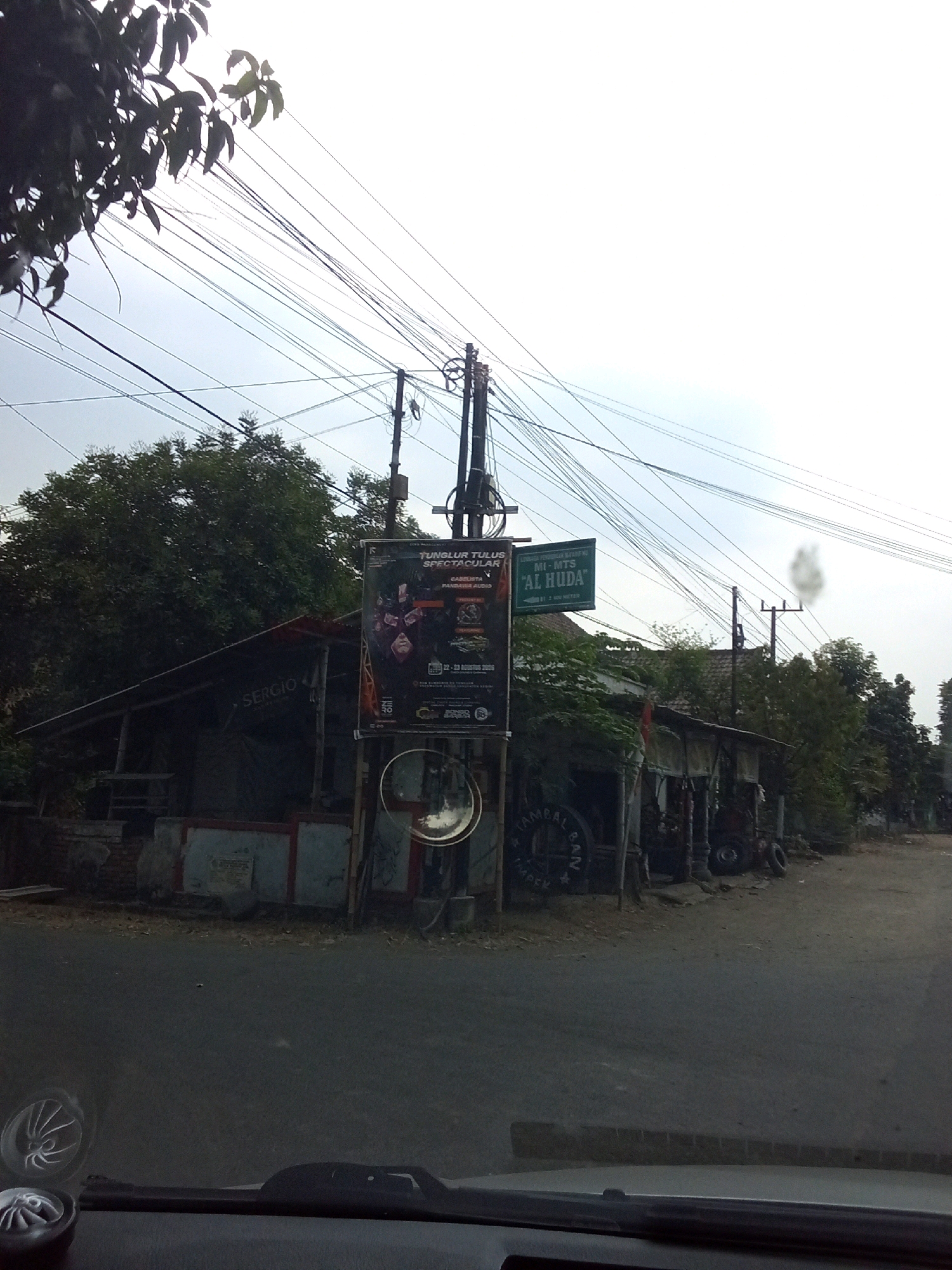 The image depicts a street scene in the city of Badas, Indonesia, under an overcast sky. In the foreground, the dashboard of a vehicle is visible, with part of a windshield wiper and a decorative cap on the dashboard. The road surface appears to be asphalt.
Further back, a building with a dark, sloping roof is situated on the left side of the image. The name "SERGIO" is visible on a sign attached to the building. A large poster is displayed on a pole in front of the building, featuring text in Indonesian that appears to be an advertisement for an event or performance. Below the poster, a circular convex mirror is mounted on a pole.
To the right of the poster, a green sign with white lettering reads "MI-MTS 'AL HUDA'". Further into the scene, more buildings and trees are visible, along with utility poles and numerous overhead electrical wires that create a dense network against the sky. There are also some parked vehicles and what appear to be storage or workshop structures. The overall impression is that of a modest commercial or residential area in a town or city.