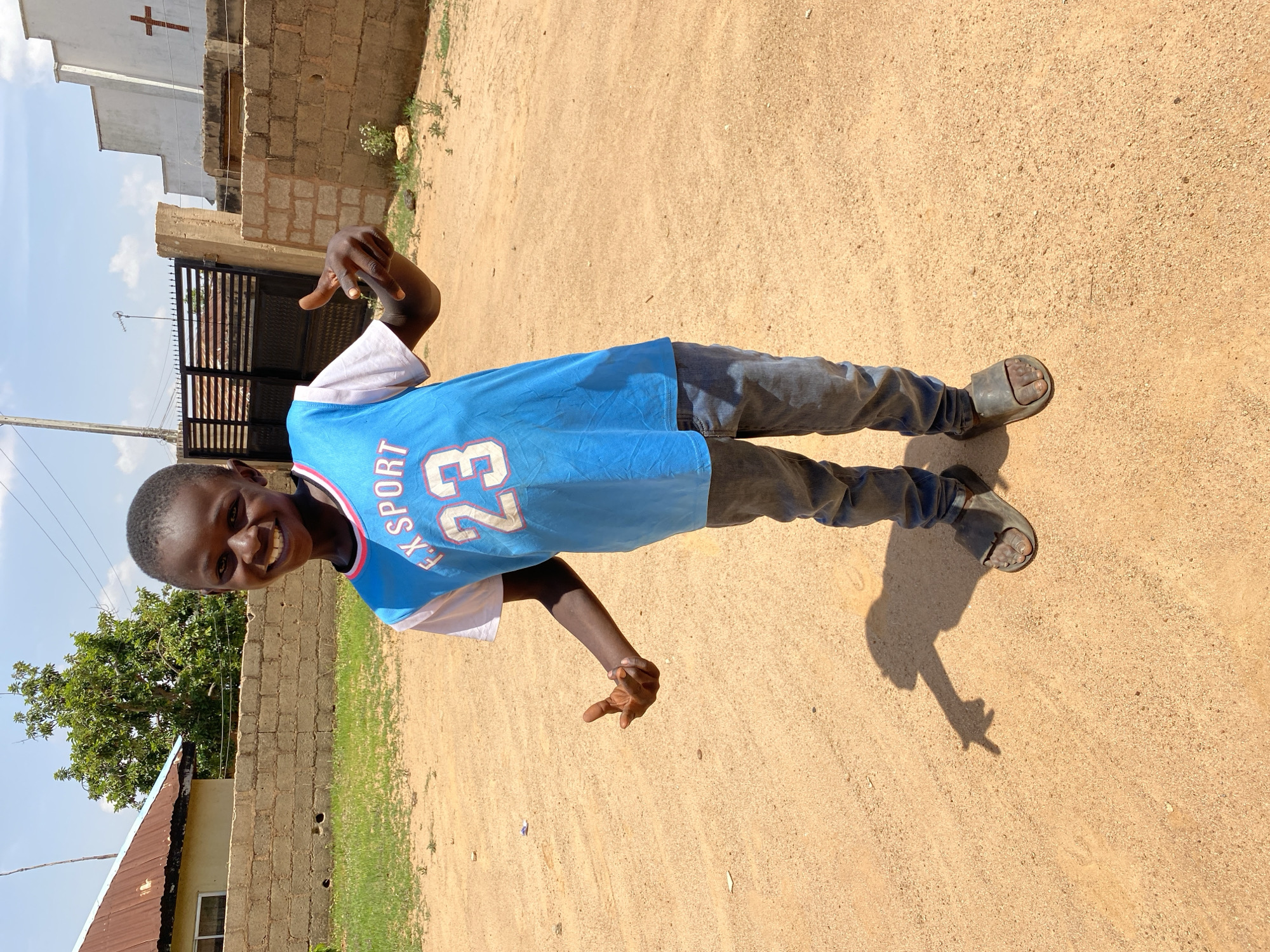 A smiling child is standing on a dirt road in Bukuru, Nigeria. The child is wearing a blue jersey with the number 23 on it, jeans, and sandals. They are making hand gestures with both hands. Behind the child is a brick wall, a building, a tree, and a metal gate. In the background, there is a building with a cross on top. The sky is cloudy.