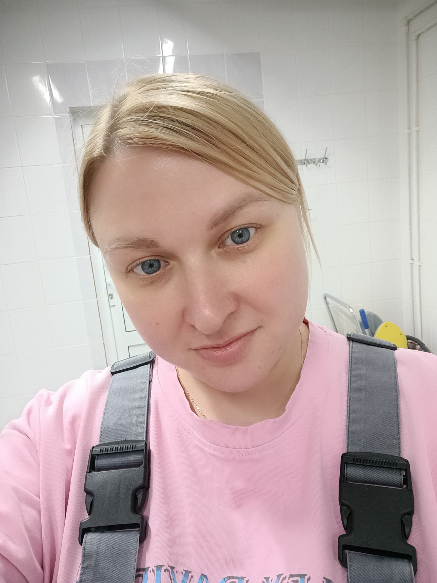 A medium close-up selfie shows a woman with blonde hair pulled back, featuring blue eyes and a fair complexion. She is looking directly at the camera with a subtle, closed-mouth smile. She wears a pink crew-neck t-shirt and grey overalls, identified by the prominent grey strap with a black plastic buckle crossing her chest and over her right shoulder. A thin gold chain is visible around her neck.
The background consists of white tiled walls, suggesting an indoor, utilitarian environment such as a kitchen, bathroom, or service area. To the left, a white door or partition is partially visible. On the right side of the frame, behind her shoulder, white pipes run along the wall, and elements resembling cleaning equipment or a cart with yellow and blue parts are discernible. A small rack with hooks is also mounted on the tiled wall. The lighting appears artificial and even.
Some flyaway hairs are visible around her part. Partially obscured text is visible on the bottom of her pink t-shirt, but it is not fully legible, showing fragments like "YVA" and "VE". The image does not provide cues for the time of day or weather.