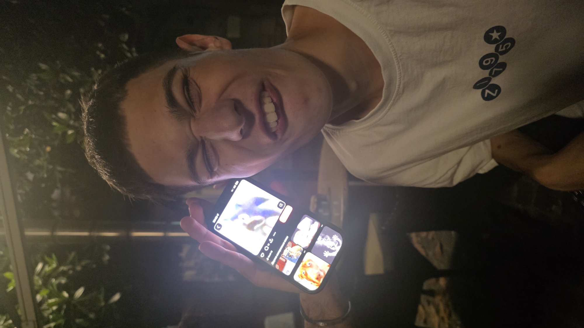 A medium shot depicts a young male holding a lit smartphone in front of him. The phone screen displays a social media app with thumbnails of various videos. He is wearing a white T-shirt with a row of blue, circle-enclosed figures on the shoulder. The setting appears to be outdoors at night, possibly in Podgorica, Montenegro, though specific identifiers are absent. The lighting is dim, and the background is dark with vague shapes suggesting trees or foliage. The man's face is slightly contorted, hinting at a grimace or strained expression.