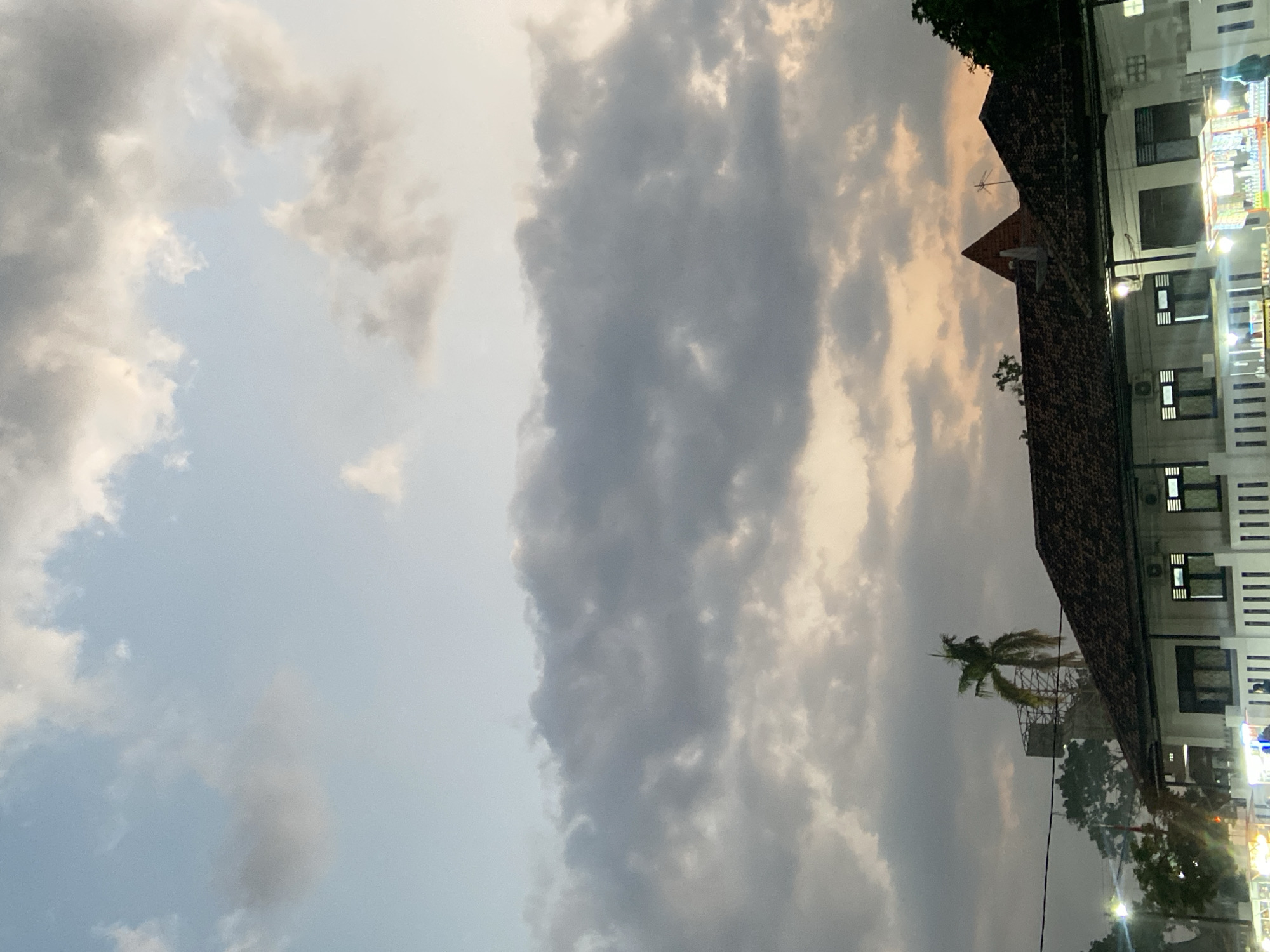 The image shows a dusky sky filled with a mixture of light and dark clouds over a low-rise building in Kecamatan Pati, Indonesia. The building has a tiled roof and features several windows and air conditioning units. In the foreground, there are some street stalls with bright lights, suggesting a marketplace or commercial area. A single palm tree stands tall to the left of the building, with a large, unfinished structure behind it. The overall scene captures a moment at dusk, with the sky displaying a gradient of colors from light blue to warmer tones.