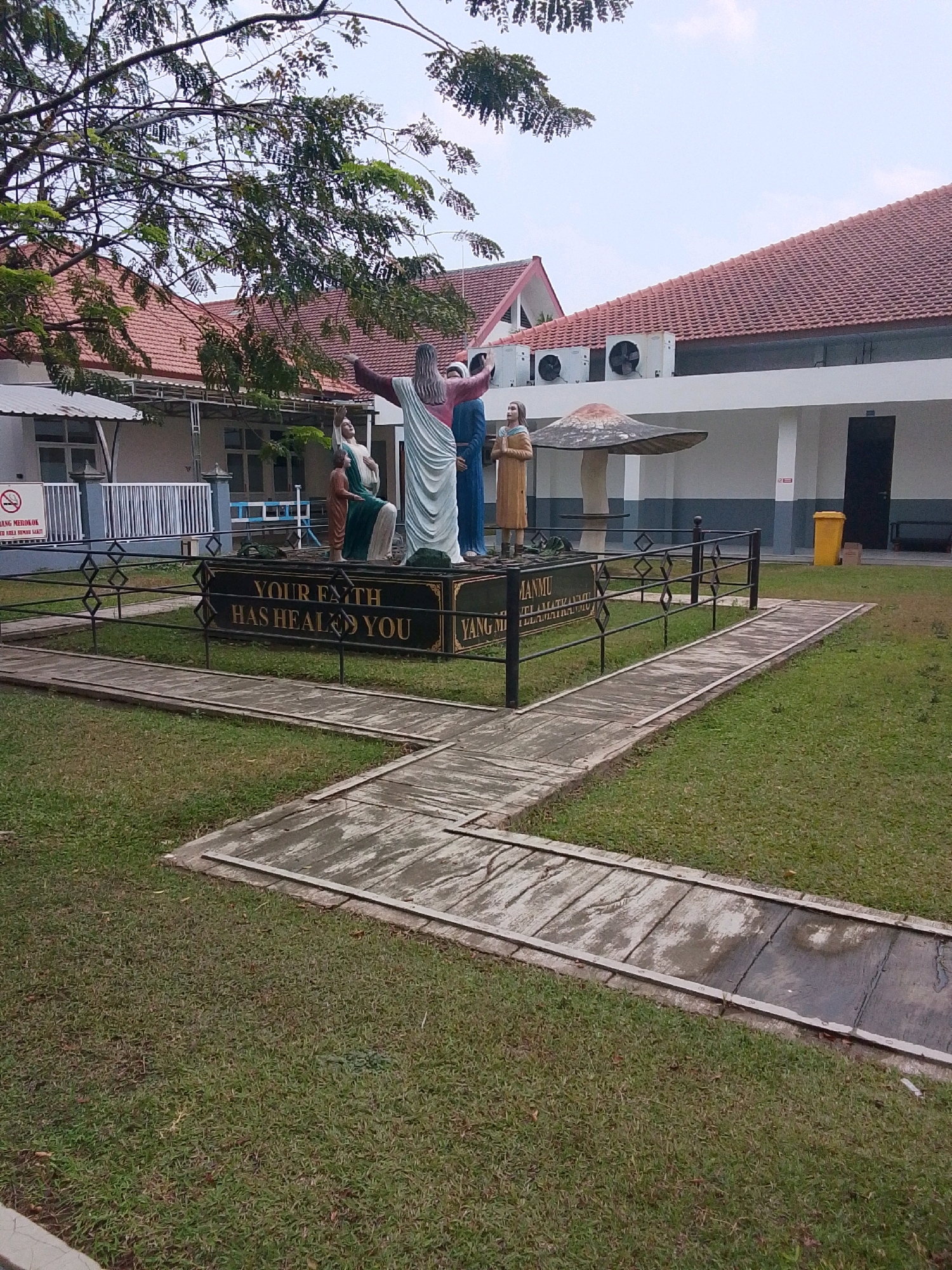 The image depicts a courtyard scene in front of a building with a red-tiled roof, located in Mojowarno, Indonesia. In the foreground, a statue depicting Jesus with outstretched arms stands center, with several other figures and a mushroom-shaped sculpture behind it. A black metal fence encloses the statue, and a pathway made of concrete slabs winds through a grassy area. The background features the building, with visible air conditioning units on the roof and a yellow trash can near a dark doorway. A sign with a red circle and slash indicating "no smoking" is visible on the left side of the frame.