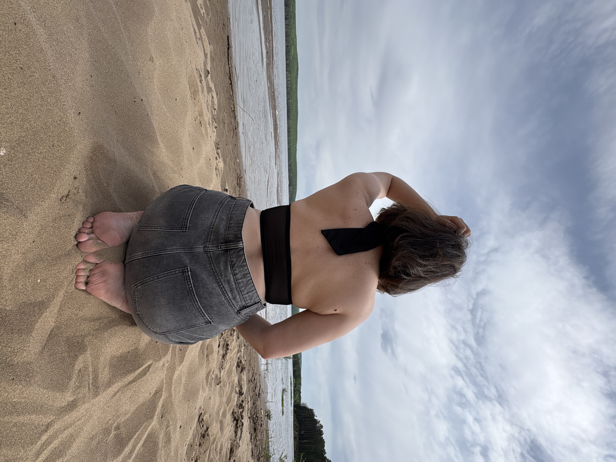A person is kneeling on a sandy beach. They are wearing gray jeans and a black halter top. Their back is facing the viewer. The water is calm and lined with trees in the background. The sky is cloudy. This scene is located in the city of Chaikovsky, Russia.