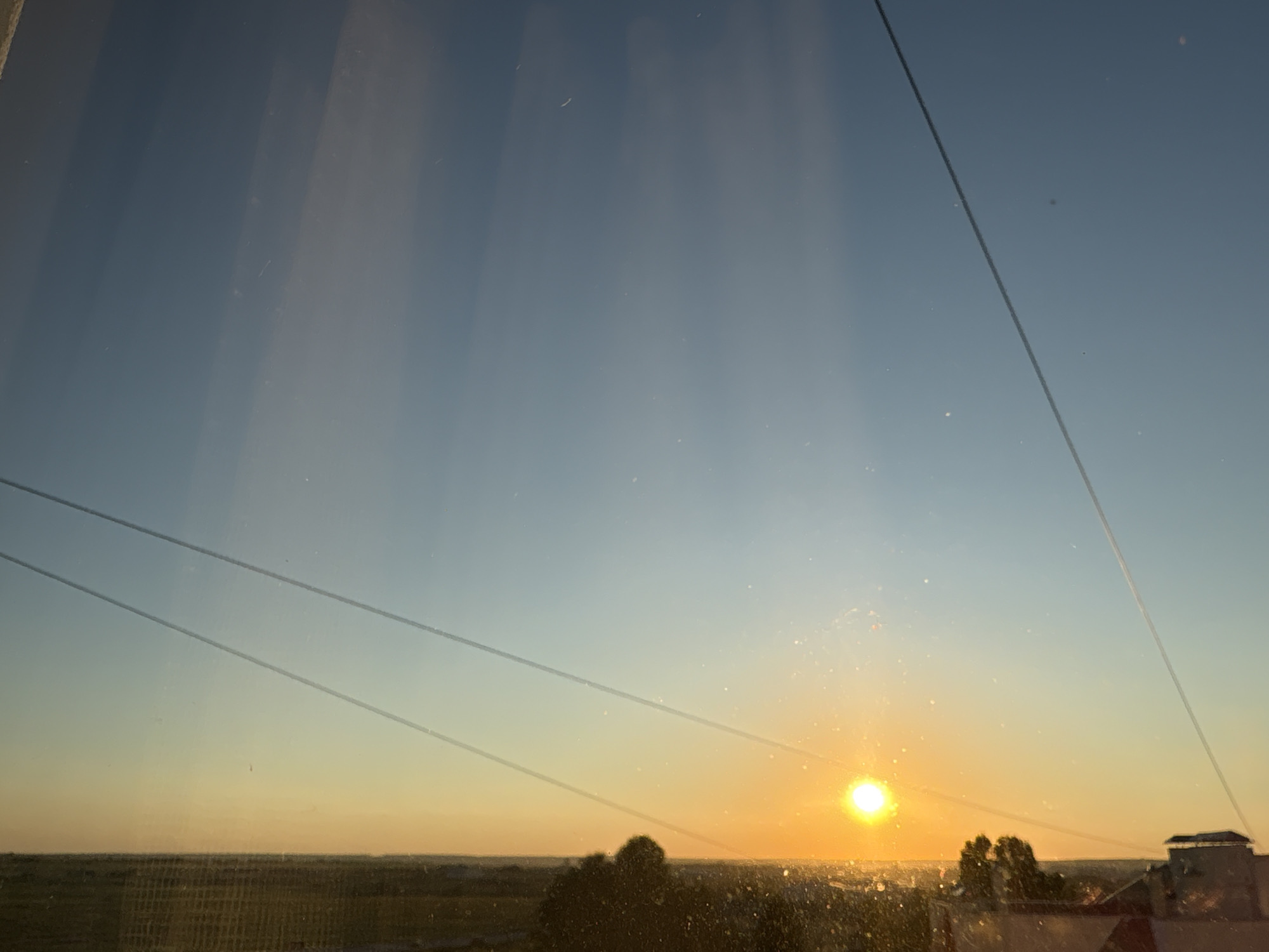 This image captures a sunset scene as viewed through a window, with distinct horizontal lines likely caused by the window panes or screen. The sky transitions from a pale blue at the top to a warm orange and yellow near the horizon, indicating the sun is low in the sky. The sun itself is a bright, concentrated orb of light on the right side of the frame, casting a golden glow. In the foreground, a blurred landscape is visible, featuring silhouetted trees and some low-rise buildings. The overall impression is that of an urban or semi-urban environment at dusk. The location is identified as Засечное, Russia, suggesting this view is from a residential or elevated vantage point within that locality. There are no people or animals visible in the frame, and the focus is on the natural light and the broad strokes of the landscape.