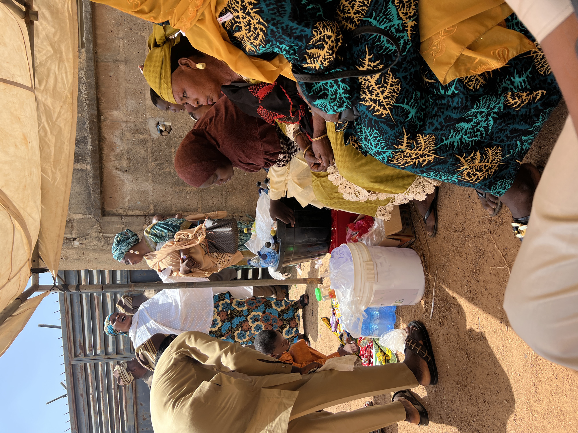 The image shows a gathering of people, primarily women dressed in colorful traditional attire, in Rigasa, Nigeria. They are sitting and standing around a dirt ground under a makeshift tent. A young boy in an orange outfit is present amidst plastic bottles and containers. A metal gate structure is visible in the background. The setting suggests a social or community event.