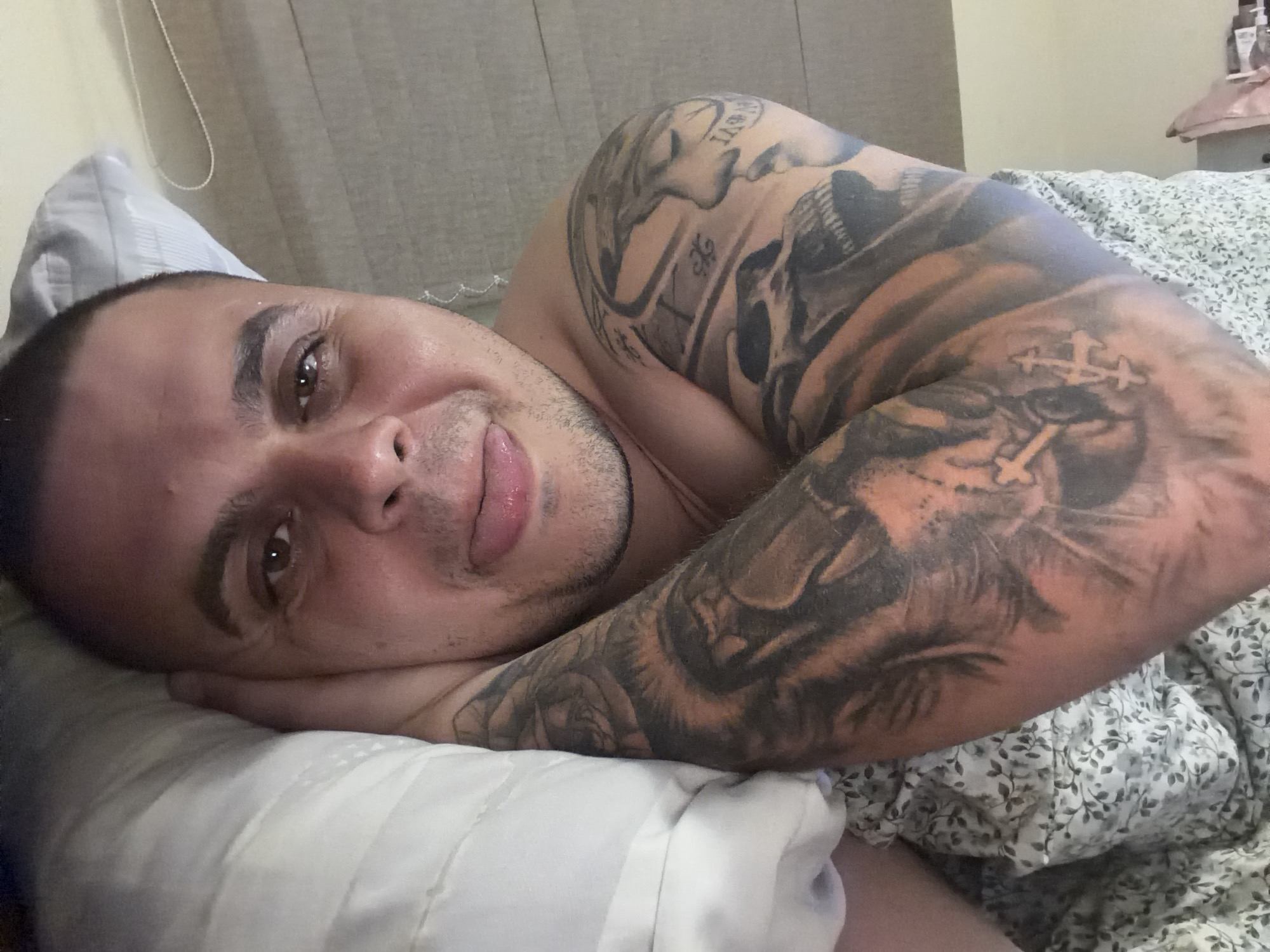 A man is depicted lying in bed indoors, likely in a bedroom in Shumen, Bulgaria. He is seen from the chest up, resting his head and right arm on a white pillow. He has a shaved head, light stubble on his face, and dark brown eyes. He looks directly at the camera with a calm expression and a gentle, slight smile.
His right arm and shoulder are extensively covered in tattoos. Notable elements of the tattoos include two stylized crosses on his forearm, a detailed image resembling a lion's head or a face, and some indistinct text or Roman numerals on his upper arm. A patterned duvet or blanket with a gray and white leaf design covers the lower part of his body. The background features a plain light-colored headboard or wall, with a thin white string or cord visible. The lighting appears soft and indirect.