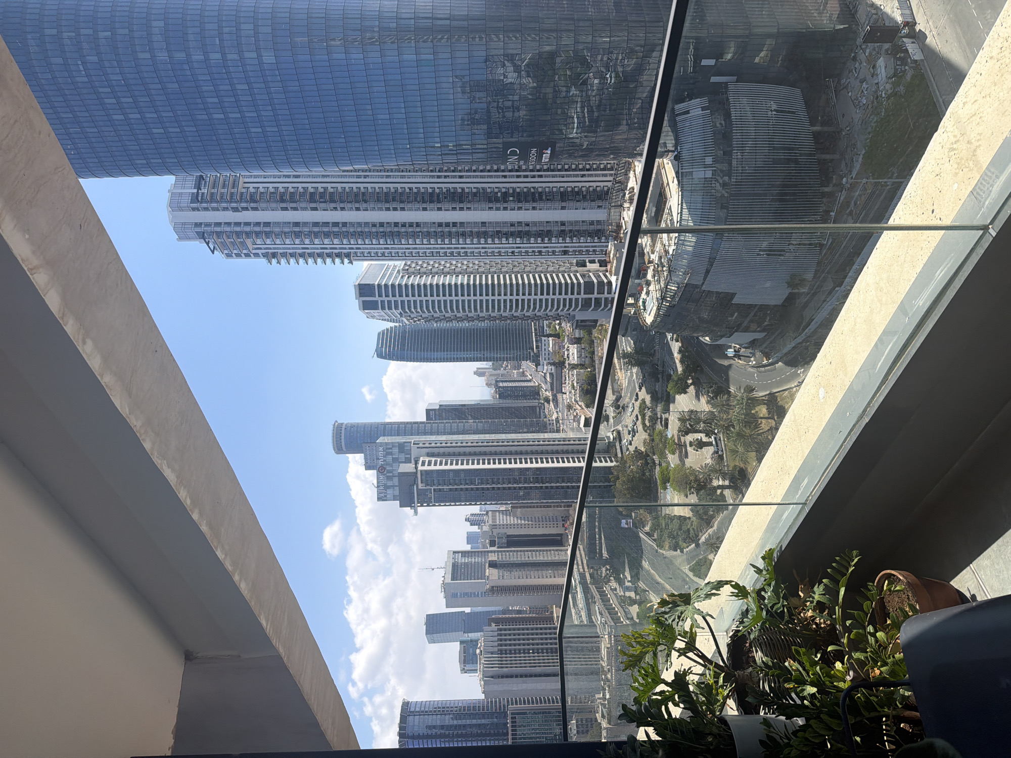 The image depicts a view from a high-rise balcony overlooking the city of Tel Aviv, Israel. Numerous skyscrapers are visible, with blue skies dotted with clouds. The balcony has a glass railing and several potted plants. The scene features a reflection of the cityscape in the glass railing. The perspective suggests a substantial height.