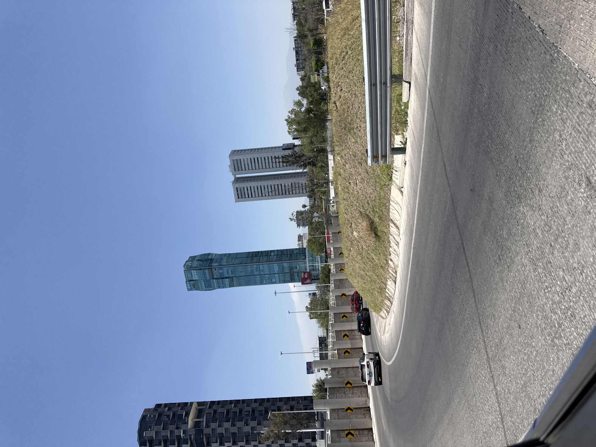 The image captures a vibrant, sunlit urban landscape, likely taken from a vehicle on a multi-lane highway. The road, featuring both asphalt and textured concrete sections, curves to the right in the foreground, with a white dividing line visible. A silver metal guardrail runs along the left side of the road, separated by a patch of dry grass.
Several cars are driving on the road, including a white car with a black roof, followed by a dark-colored vehicle, and a red car further behind. Along the side of the road, concrete barriers with prominent yellow and black chevron markings are visible, guiding traffic.
The background reveals a modern city skyline under a clear, bright blue sky, indicating daytime. Dominating the scene are several distinct high-rise buildings. On the right, a pair of tall, rectangular, light-colored twin towers stand, with the word "inbox" faintly discernible on one of their facades. To the left, a striking, taller skyscraper with a curved, blue-tinted glass exterior and a unique architectural design rises prominently. Another large building with a dark, textured facade is partially visible on the far left. The urban environment is interspersed with trees and areas of dry, brownish grass. Streetlights line the roadway. The scene depicts a typical busy highway setting in San Bernardino Tlaxcalancingo, Mexico.